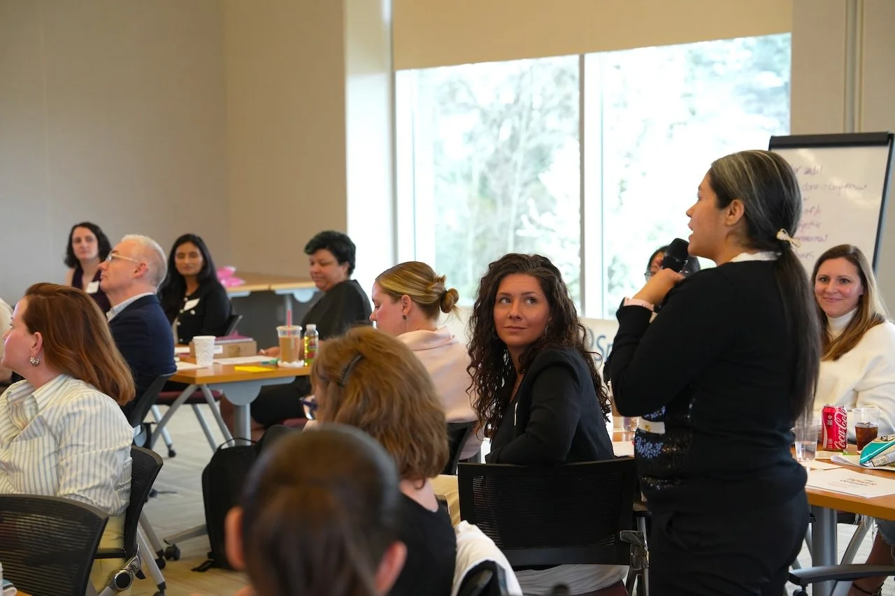 A woman standing in a conference room speaking into a microphone to an audience of diverse adults seated at tables. The room has large windows letting in natural light.