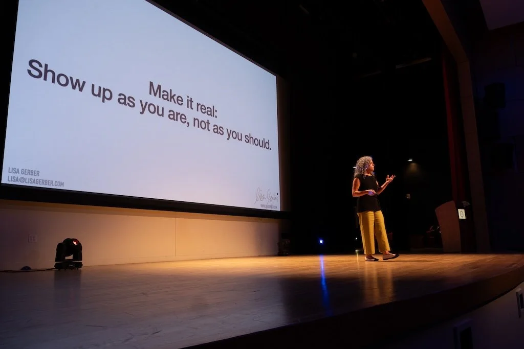 A woman with curly gray hair stands on a stage presenting to an audience. Behind her, a large screen displays a motivational quote, "Make it real: Show up as you are, not as you should," along with her name, Lisa Gerber, and her email address.