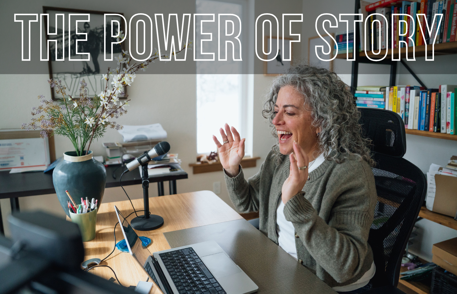 A woman with curly gray hair sitting at a desk, smiling and gesturing with her hands, in front of a microphone, with a laptop, a large vase with flowers, and a bookshelf in a well-lit room.