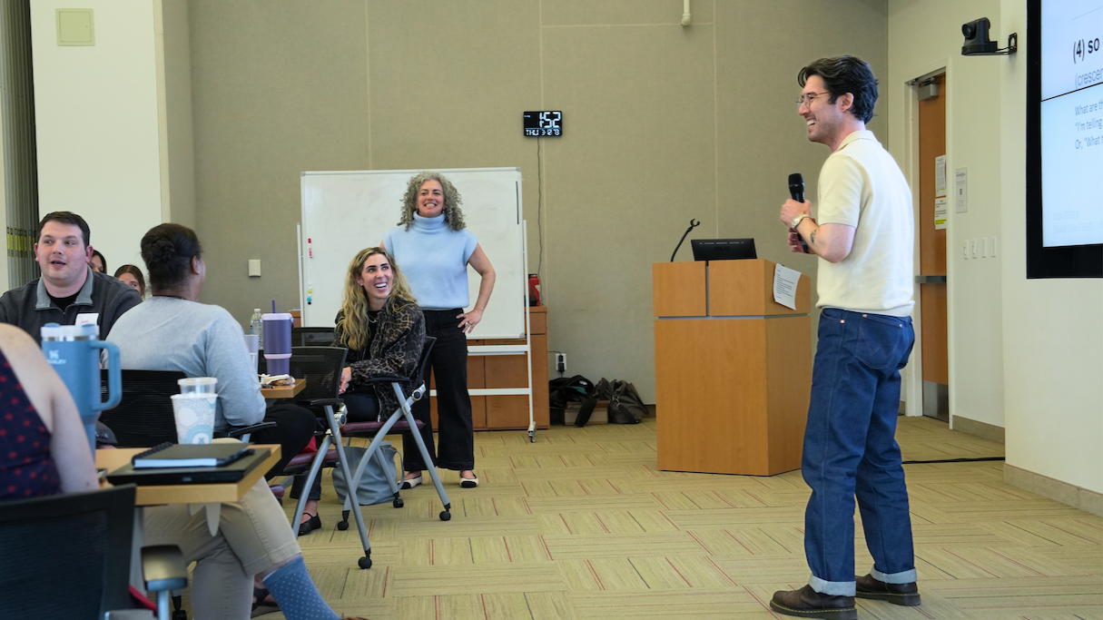 A man with glasses and dark hair holding a microphone presents to a group of people in a classroom or conference room, with some individuals smiling and laughing while seated around tables.