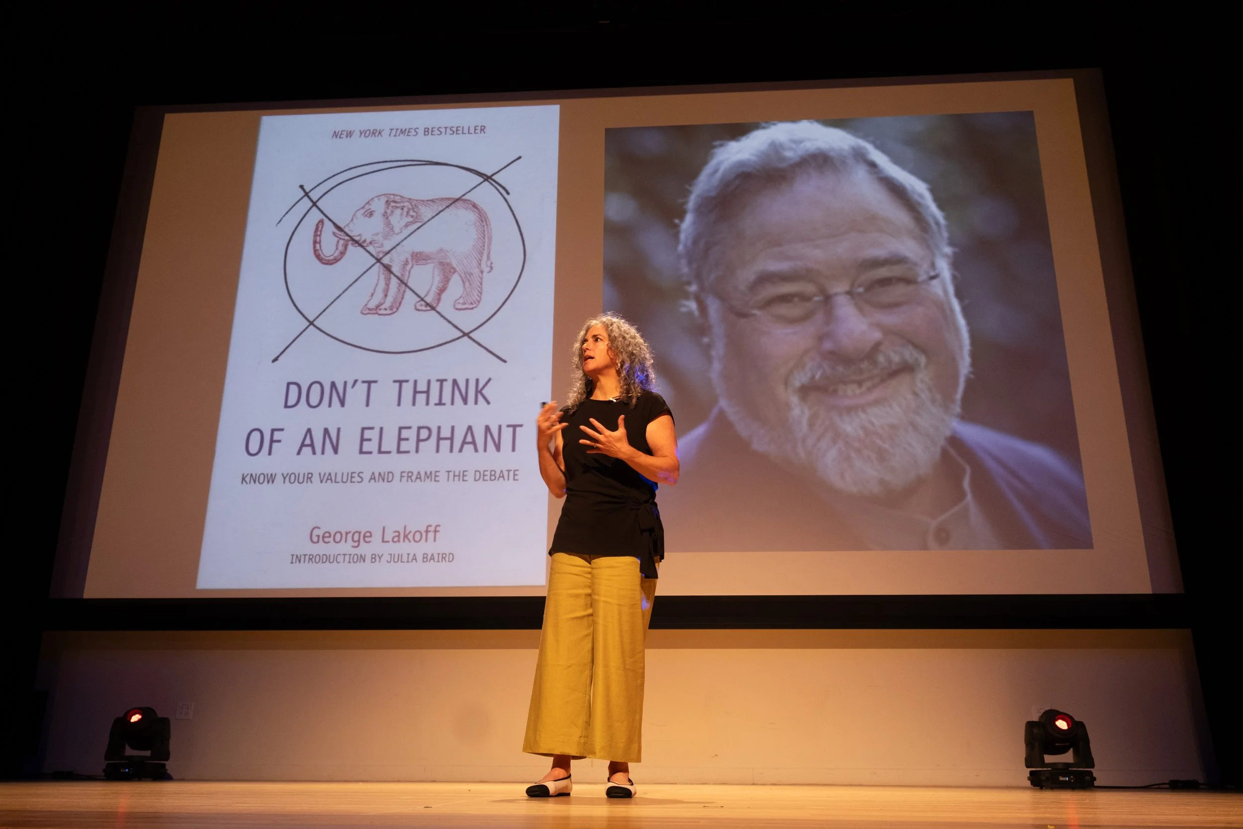 A woman with curly hair and glasses speaking on a stage, in front of a large screen displaying a book cover titled 'Don't Think of an Elephant' by George Lakoff, and a large portrait of a smiling man with glasses.