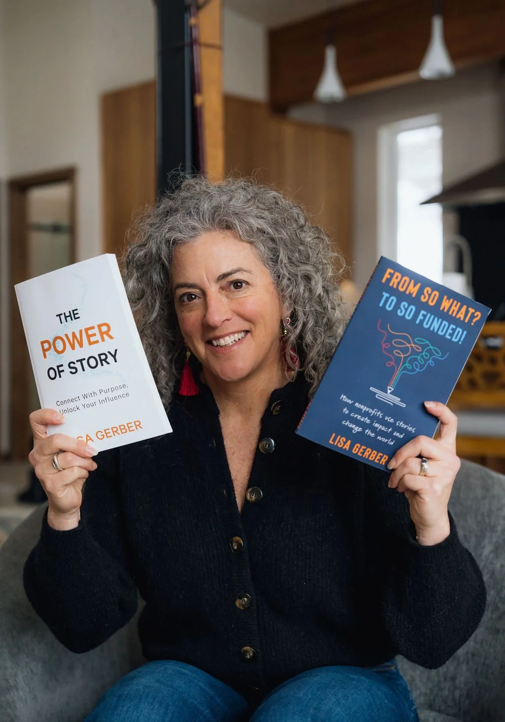 A woman with curly gray hair sitting on a gray couch, smiling, holding two books titled 'The Power of Story' and 'From So What? to So Funded!' in a cozy home setting.