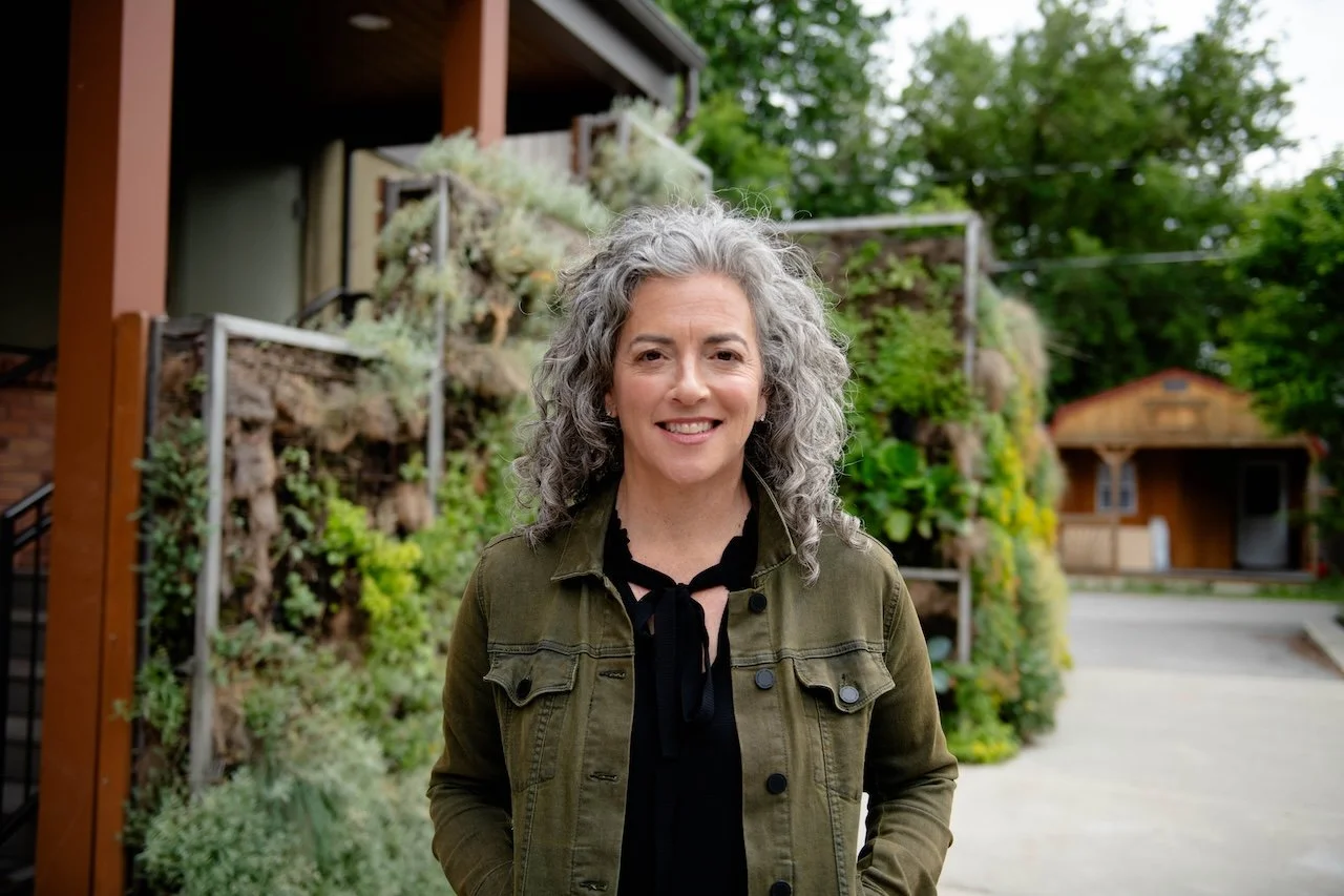 A woman with curly gray hair smiling outdoors in front of vertical garden structures and trees.