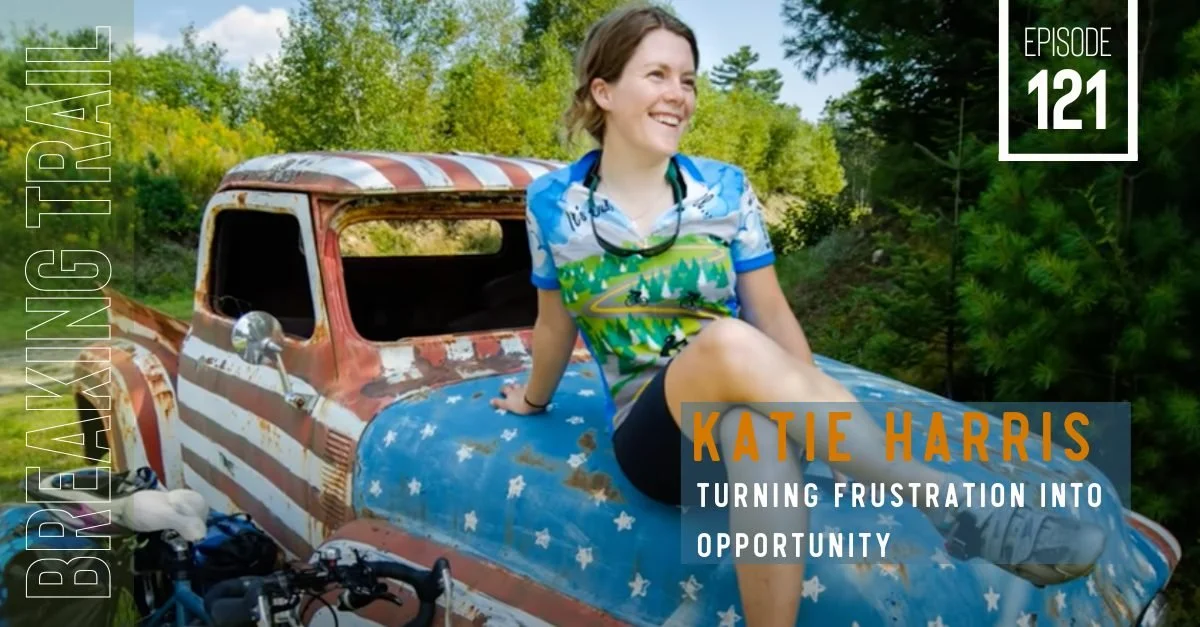 A smiling woman sitting on a vintage rusty pickup truck in a lush green outdoor setting, wearing a colorful shirt with nature scenes, with bicycles nearby.