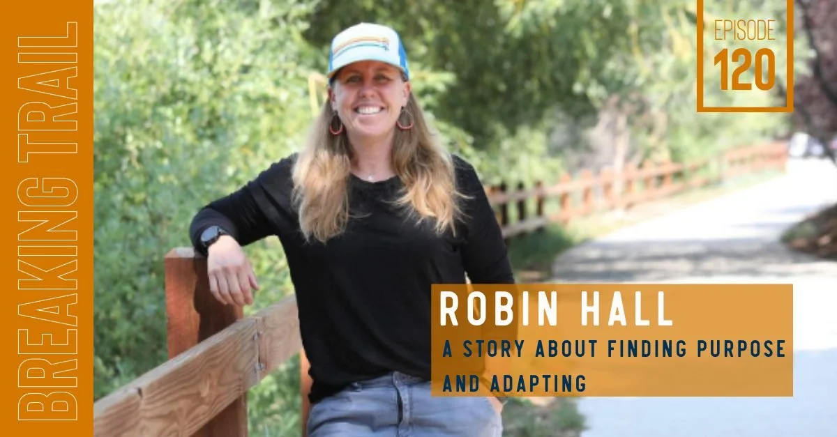 Woman smiling and leaning on a wooden railing outdoors, wearing a black shirt, blue shorts, a light blue cap, and hoop earrings, with a background of green trees and a path.