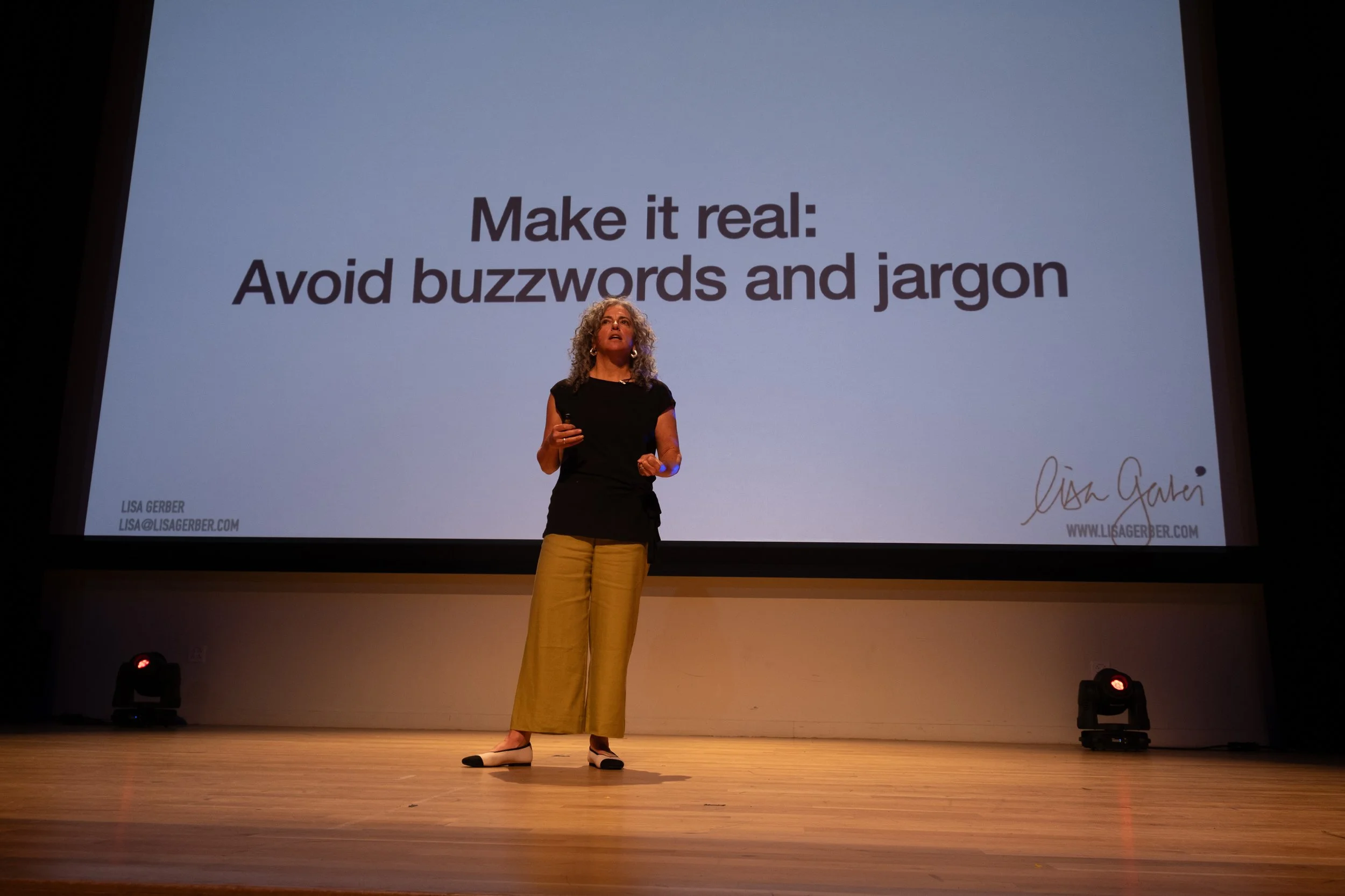 Woman giving a presentation on stage with a slide behind her that says 'Make it real: Avoid buzzwords and jargon'.