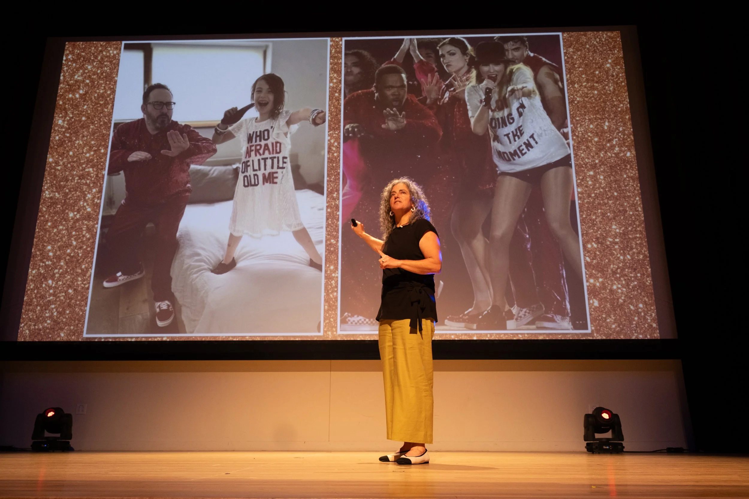 A woman with curly hair in black and yellow pants giving a presentation on a stage, with a slide behind her featuring two sets of people posing humorously. The left group shows a man and a young girl making exaggerated gestures, with the girl wearing a white shirt with red text. The right group shows a woman with a microphone and several people making energetic poses, all smiling or with expressive faces. The stage has a gold glitter border around the slide.