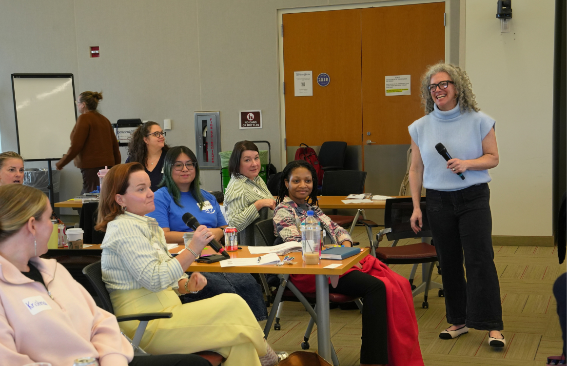 A woman with curly gray hair, wearing glasses, a light blue sleeveless turtleneck top, and black pants, stands holding a microphone in a conference room. Several seated women are listening attentively, with some taking notes or holding microphones. The room has tables, chairs, notebooks, water bottles, and a whiteboard in the background.