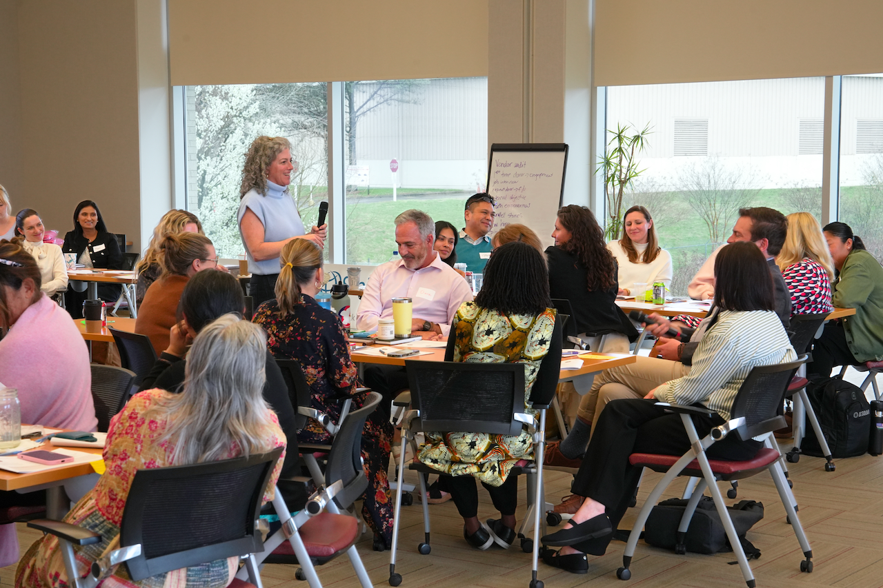 A woman speaking to a group of diverse adults during a workshop or seminar in a well-lit conference room with large windows.