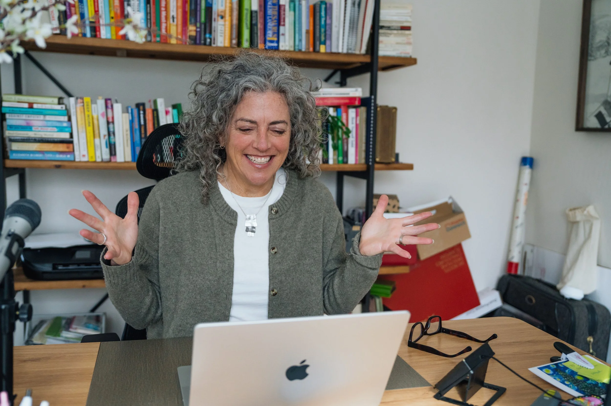 A woman with curly gray hair smiling and gesturing with her hands while sitting at a desk in a cluttered office or workspace with bookshelves, a laptop, and various office supplies.