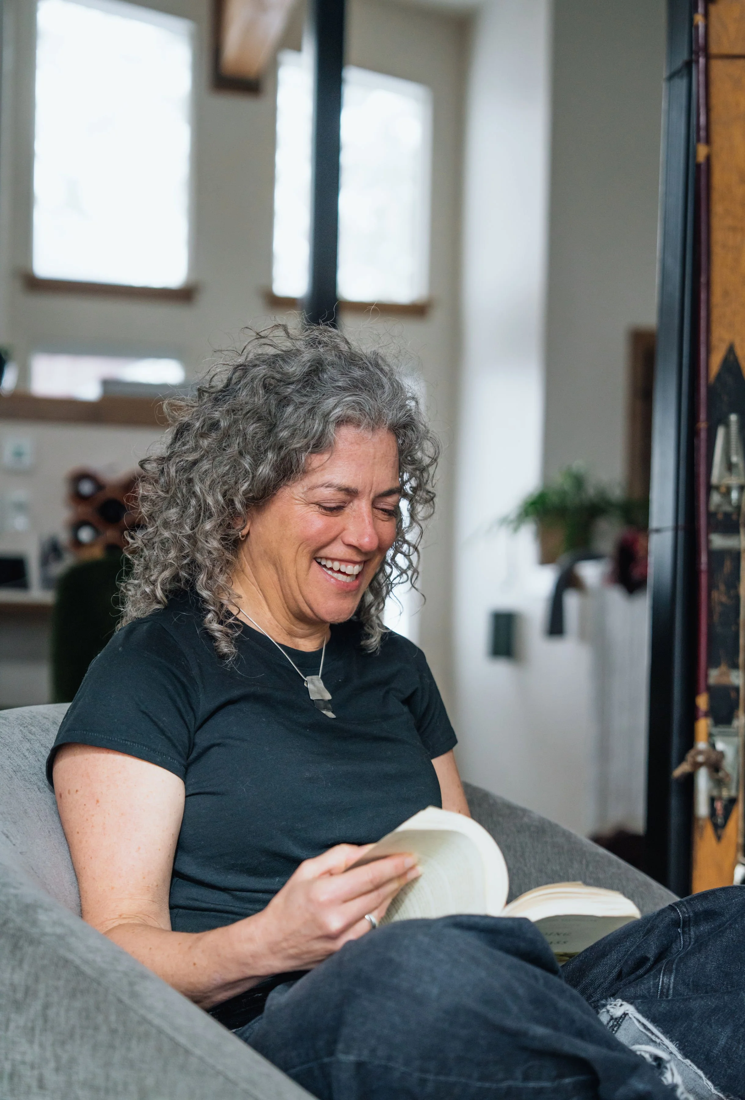 A woman with curly gray hair sitting on a gray couch, smiling, and reading a book in a living room with large windows and natural light.