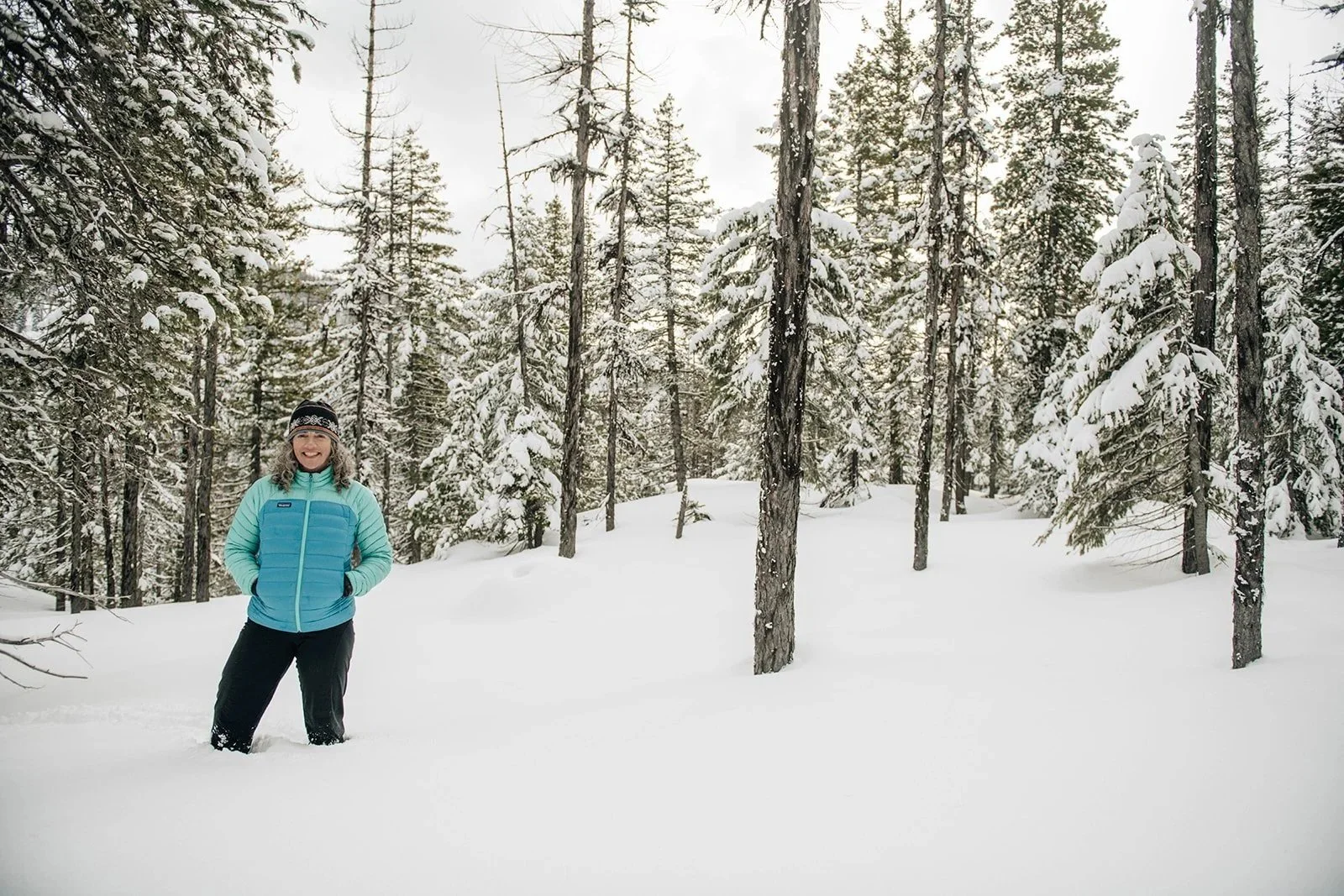 A woman dressed in a blue jacket, black pants, and a black winter hat, standing in a snowy forest with tall, snow-covered evergreen trees.