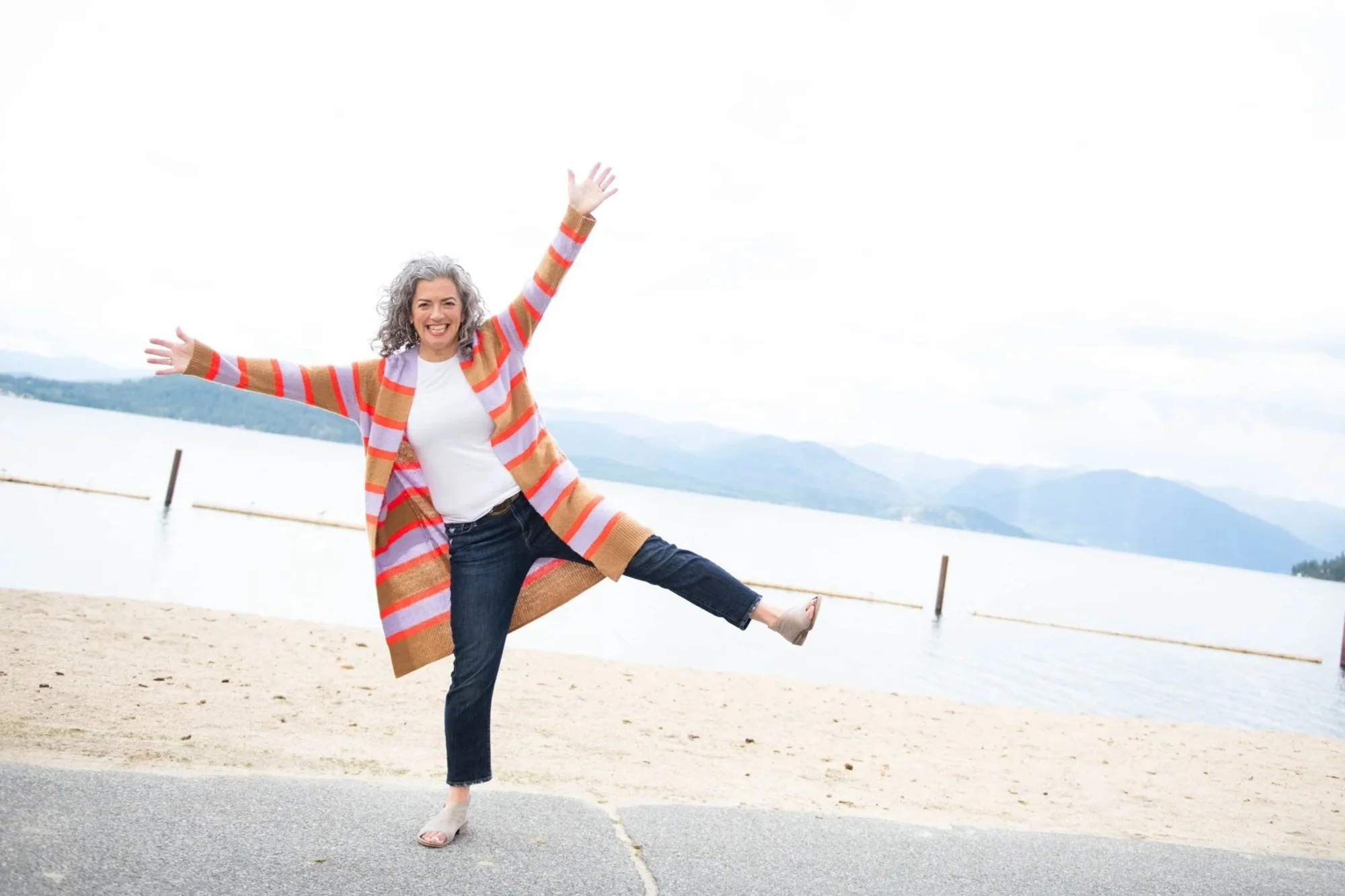 A joyful woman with curly gray hair, wearing a multi-colored striped cardigan, white shirt, and dark jeans, is standing on a beach with arms wide open and one leg lifted for a playful pose. The background features a body of water, distant mountains, and overcast sky.