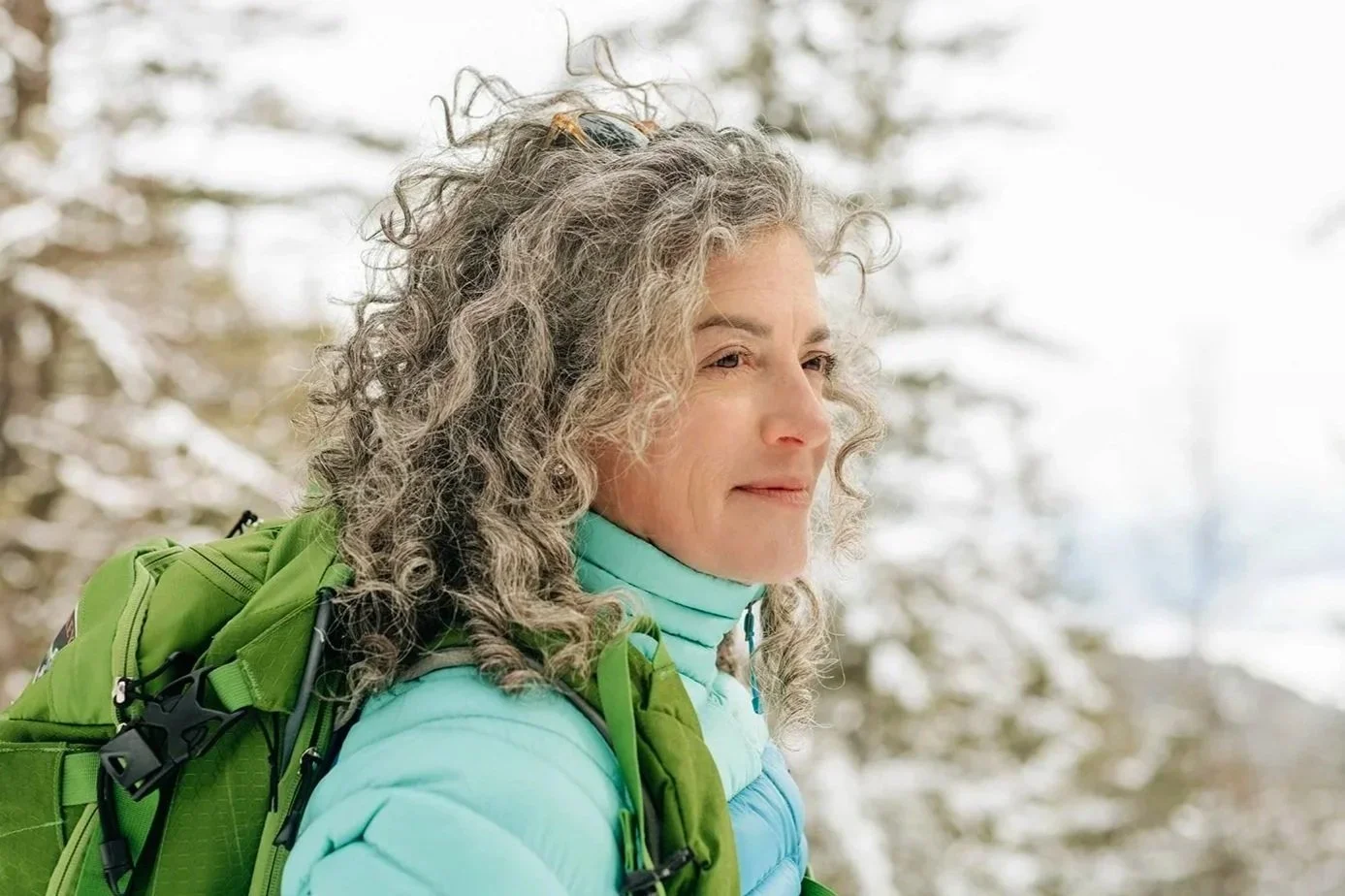 A woman with curly gray hair outdoors in a snowy forest, wearing a light blue jacket and a green backpack.