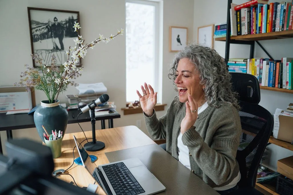A woman with curly gray hair smiling and speaking into a microphone during a video call in her home office, surrounded by a computer, books, and decorative items.