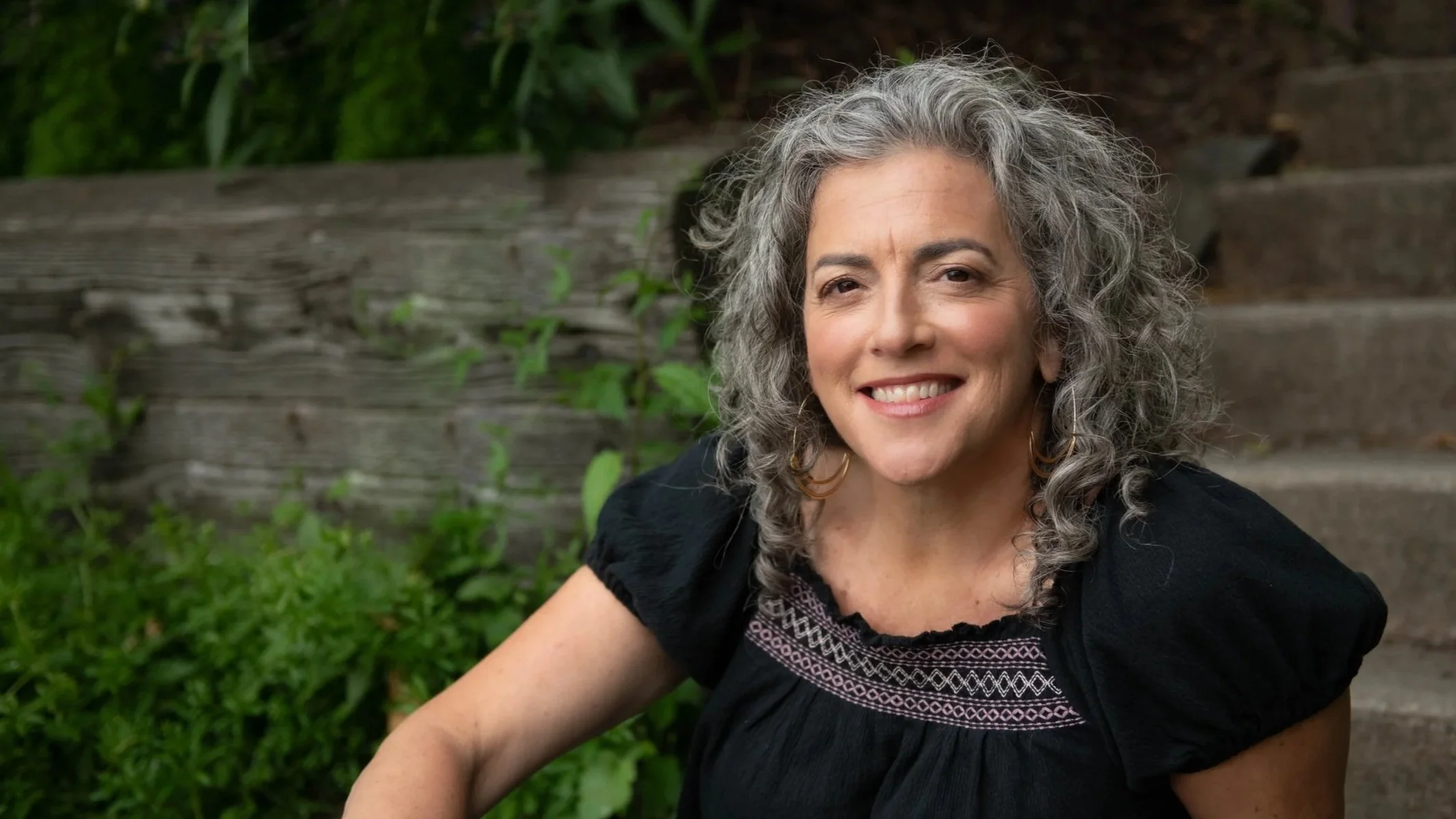 A middle-aged woman with curly gray hair sitting outdoors on wooden stairs, smiling and wearing a black top with embroidered details.