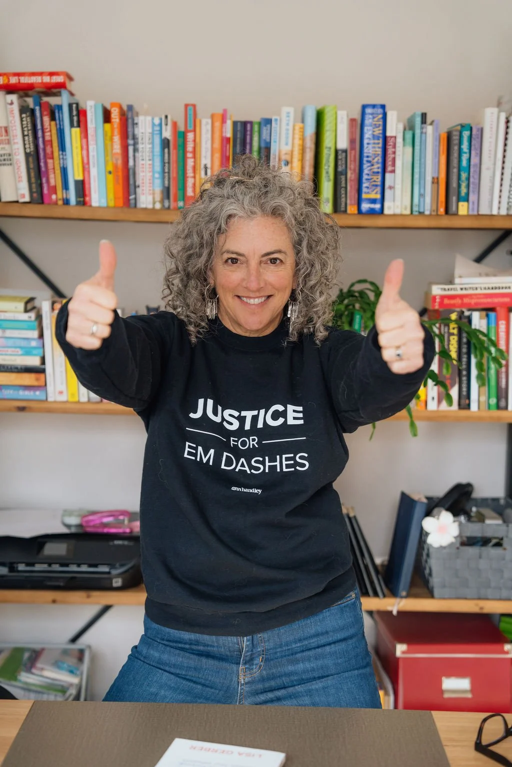 Woman with curly gray hair smiling and giving two thumbs up, wearing a black shirt with 'Justice for EM Dashes' printed on it, standing in front of bookshelves filled with colorful books.