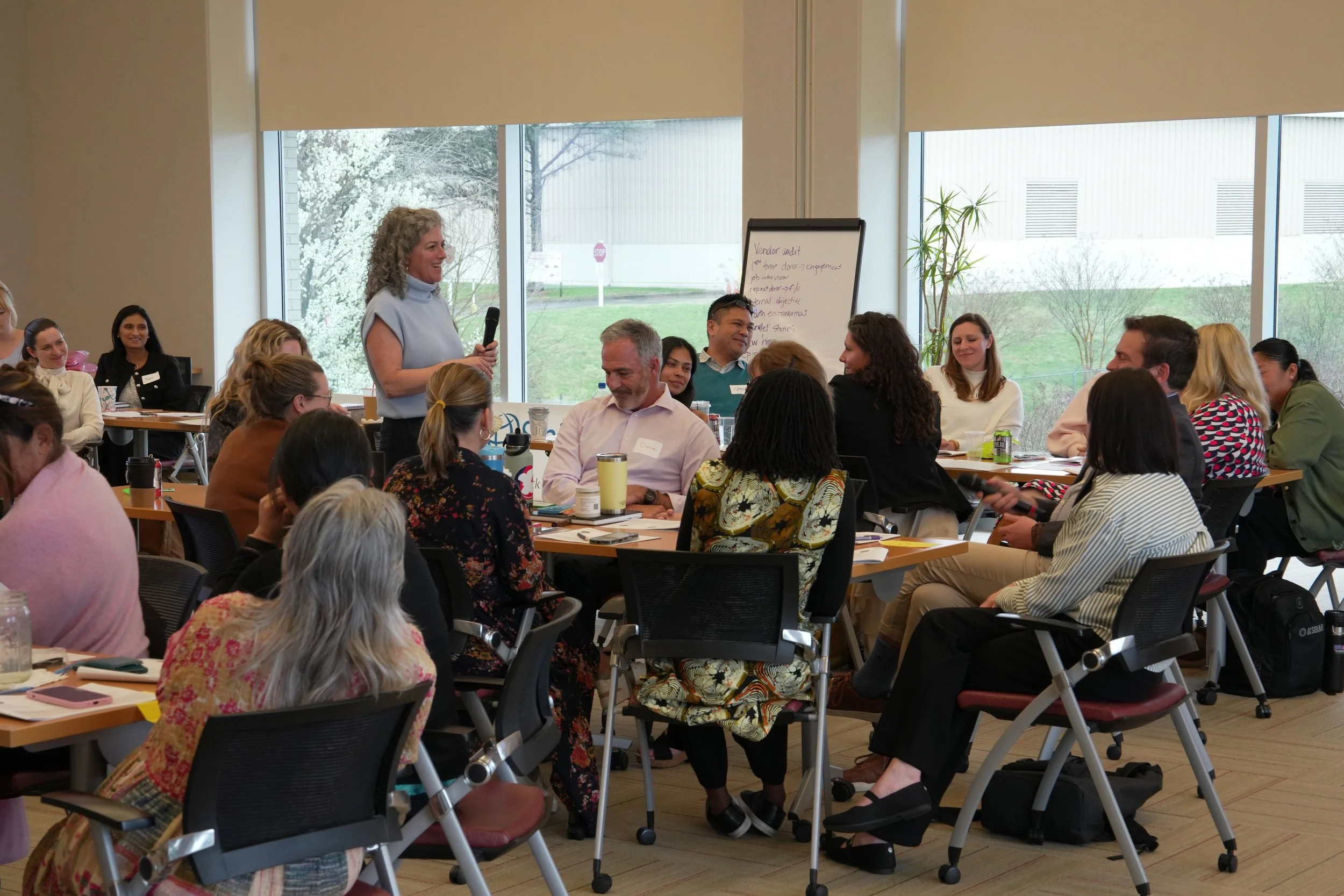 A woman with curly gray hair stands holding a microphone, speaking to a group of seated people in a conference room with large windows showing trees outside. The audience is mixed, mostly women, some taking notes or listening attentively.
