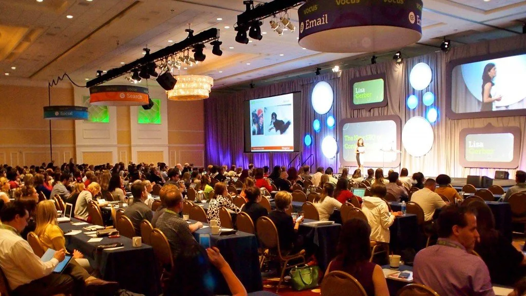 Large conference room filled with seated attendees watching a speaker on stage with multiple screens and lighting.