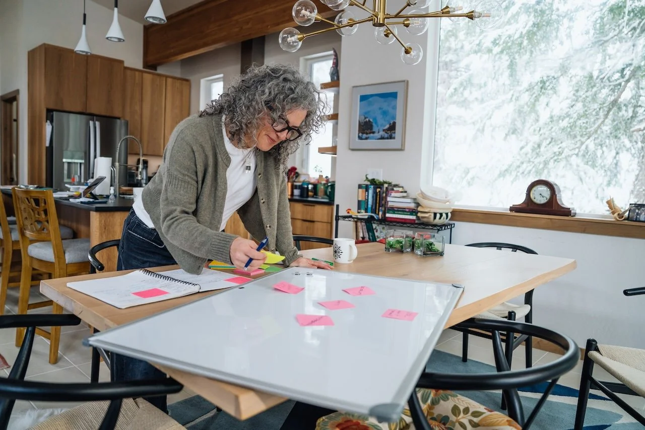 Woman with gray curly hair and glasses standing at a table, working with sticky notes and notebooks, in a bright kitchen and dining area decorated with books and a clock.