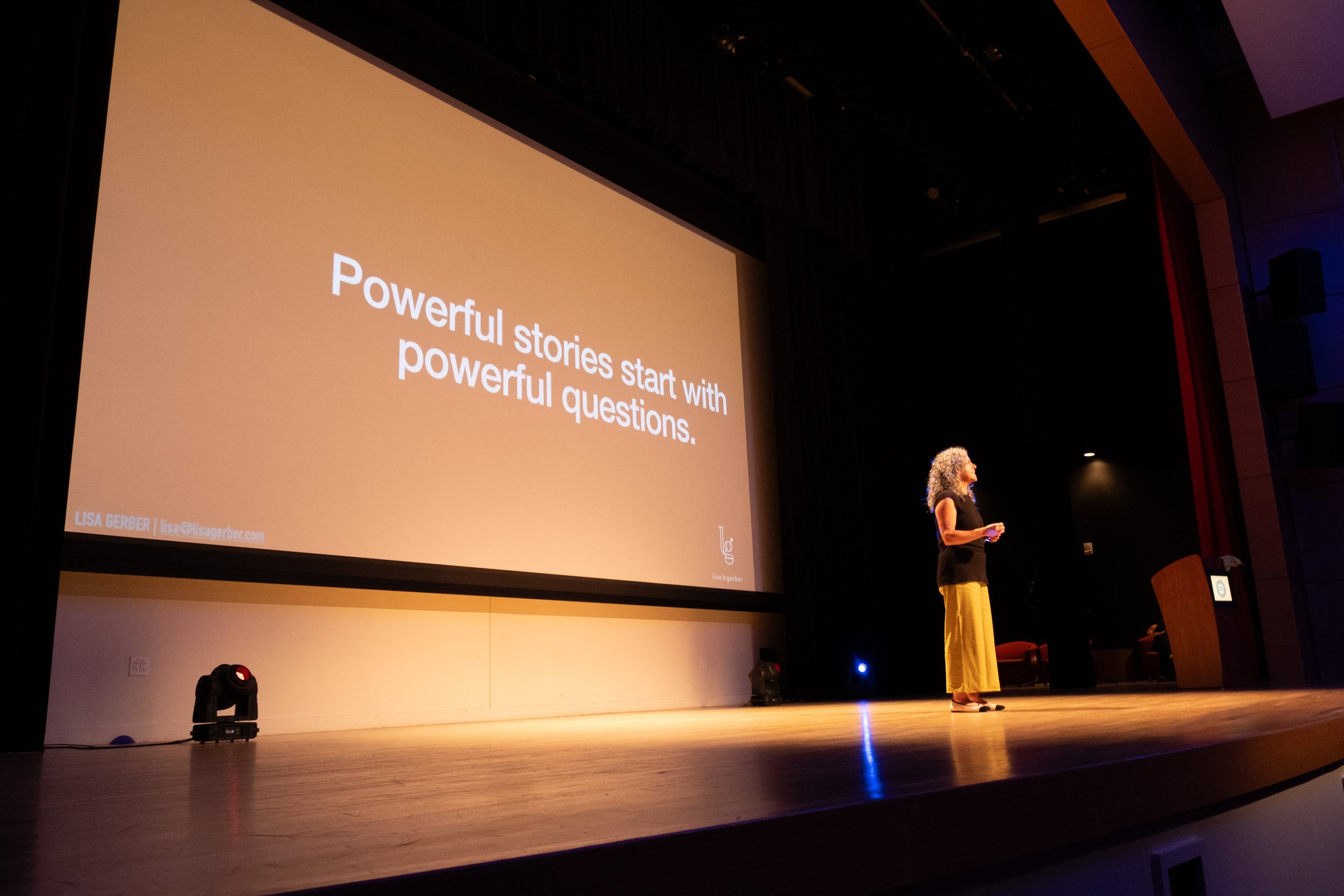 A woman standing on stage beside a large screen that displays the text "Powerful stories start with powerful questions." The stage is in a dark auditorium with a red curtain on the right side.