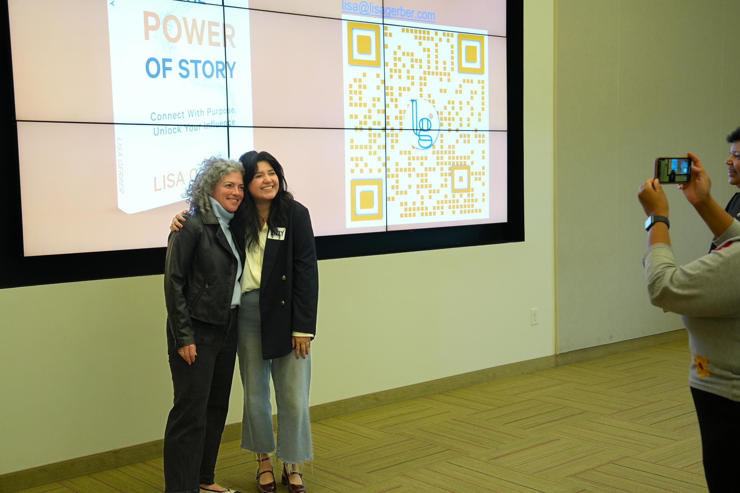 Two women standing together smiling for a photo in front of a large digital screen displaying a book titled 'Power of Story' and a QR code. The woman on the left has curly gray hair, wearing a black jacket, and the woman on the right has long dark hair, wearing a black blazer, light-colored pants, and heels. A person on the right is taking their picture with a smartphone.
