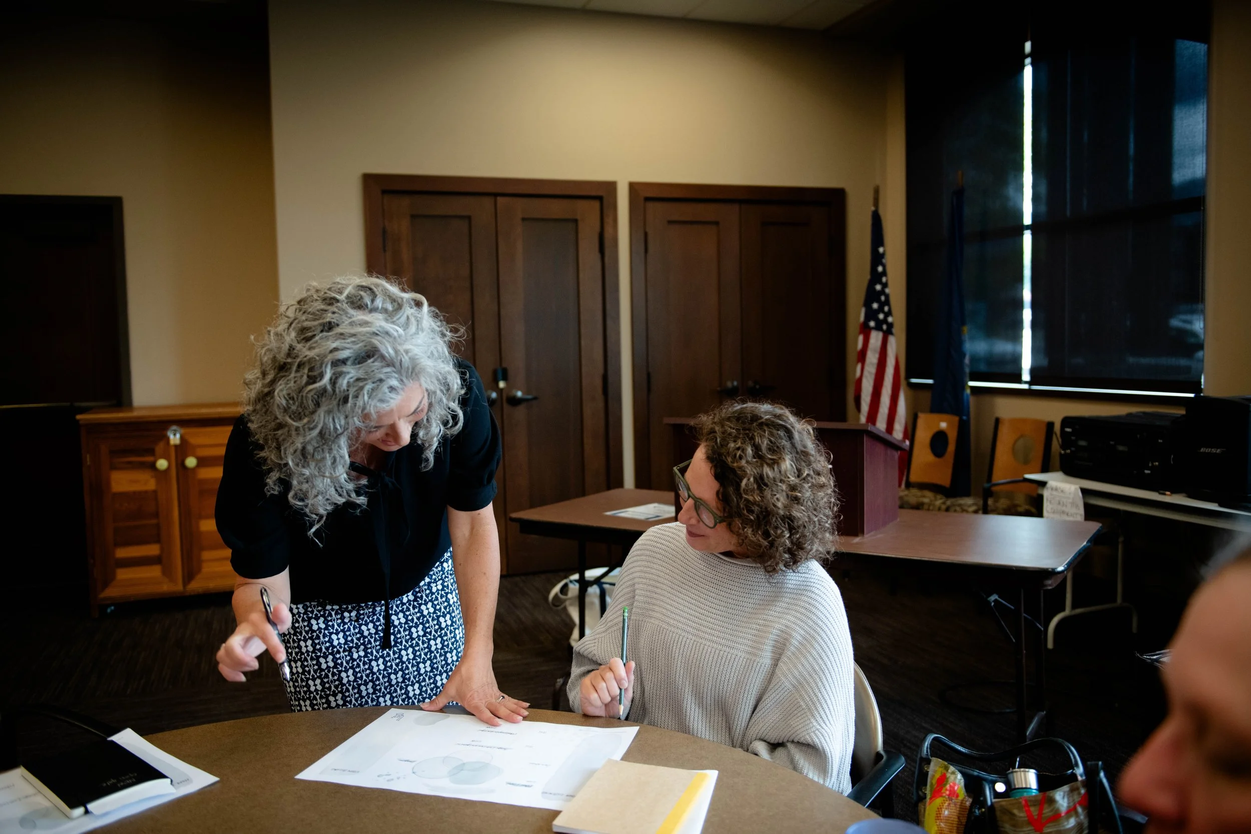 Two women are seated at a table, signing documents, while a woman with curly gray hair leans over to assist or discuss with one of the seated women in a meeting or signing event in a room with wooden doors and U.S. flags in the background.