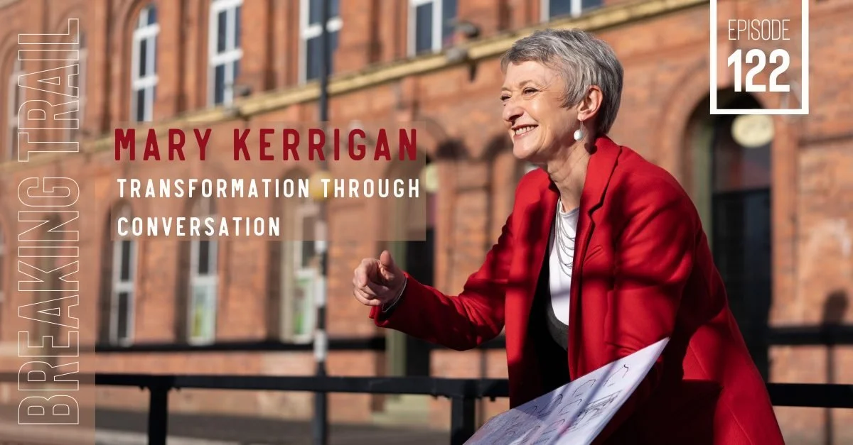 An older woman with short gray hair, wearing a red blazer and earrings, smiling and speaking outdoors in front of a brick building.