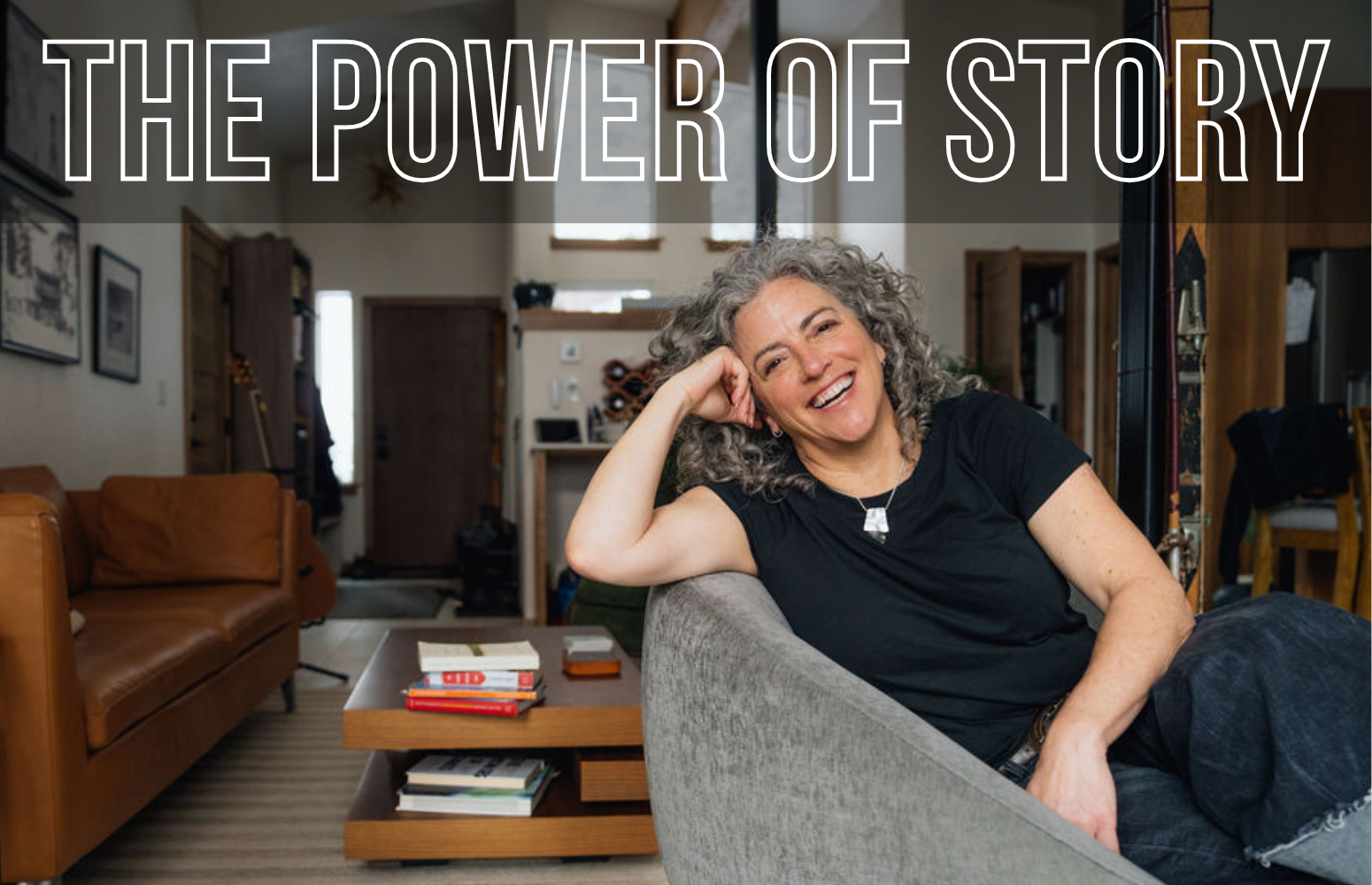 Smiling woman with gray curly hair sitting on a gray sofa in a cozy living room, with a coffee table and books in the foreground, and a tan couch, wooden furniture, and artwork on the walls in the background. Text overlay reads 'The Power of Story.'