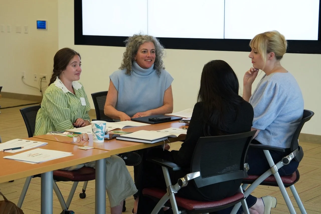 Four women having a meeting around a table in a conference room with a large screen behind them.