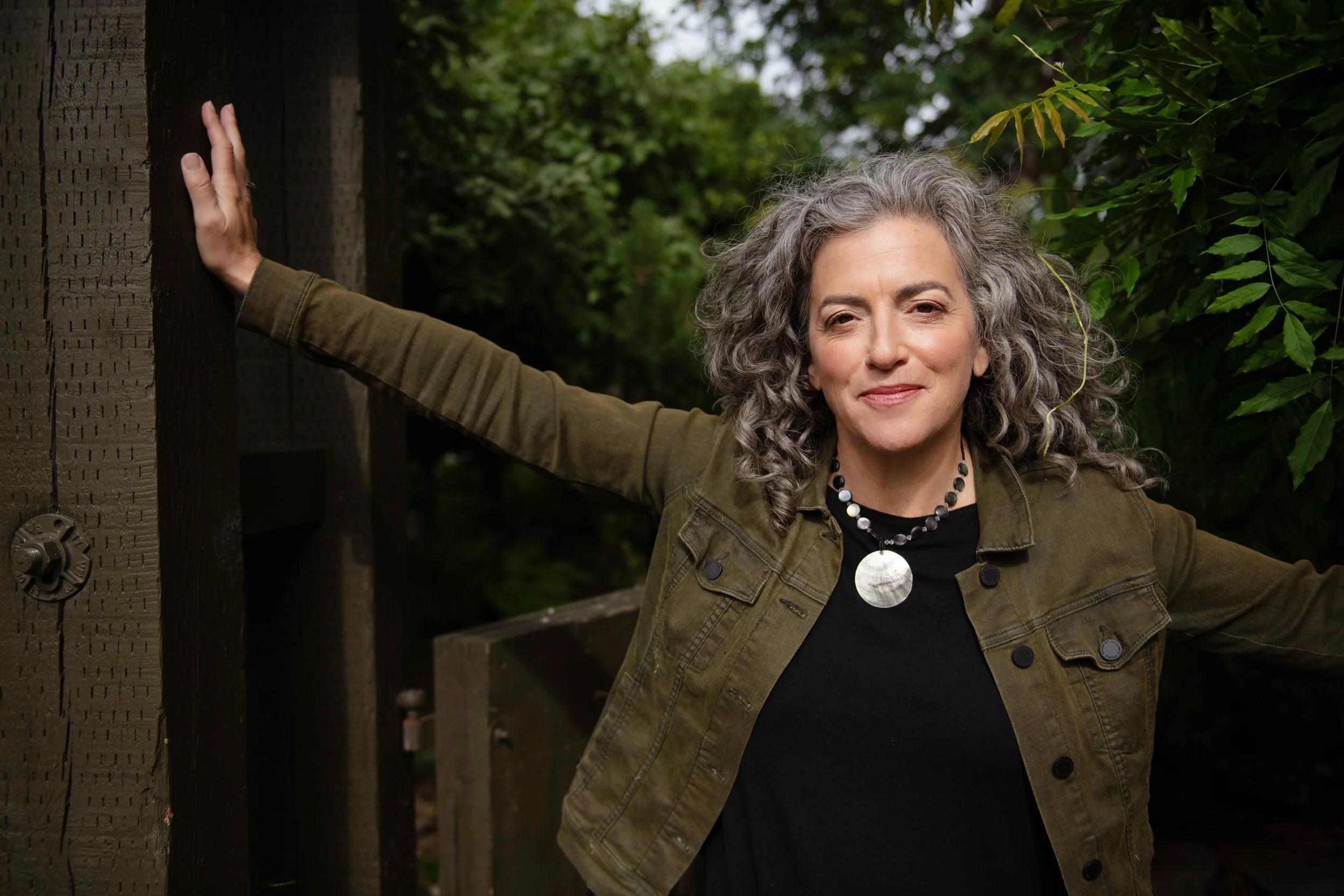 A woman with curly gray hair smiling, standing outdoors with greenery in the background, wearing a green jacket and a necklace.