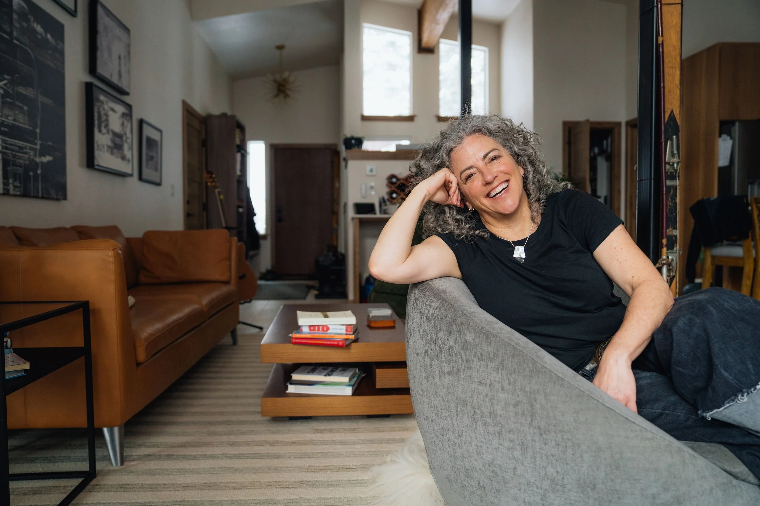 A middle-aged woman with curly gray hair smiling and leaning her head on her hand as she sits on a gray couch in a living room with natural light coming through large windows.