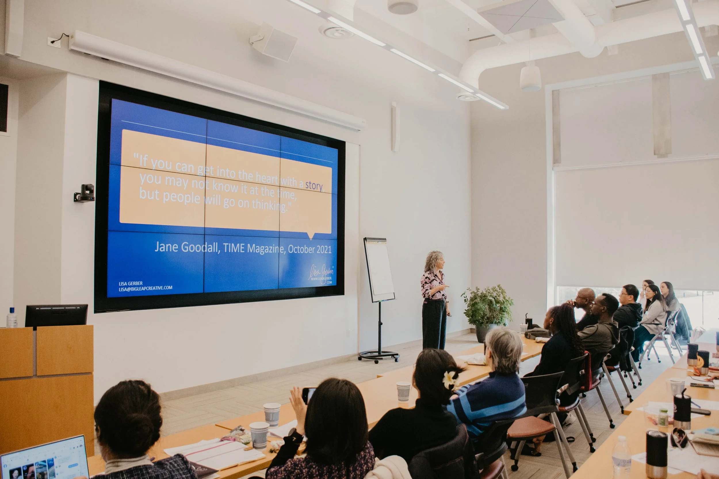 A woman giving a presentation to an audience in a conference room with a large digital screen displaying a quote by Jane Goodall. The room is filled with seated attendees, some with laptops and notebooks, and has a modern design with white walls, ceiling, and large windows.