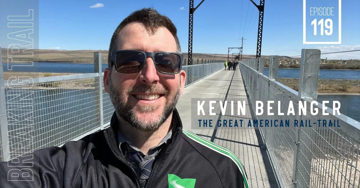 A man wearing sunglasses and a black jacket smiling on a bridge with a body of water on either side and train tracks overhead. In the background, a few people are walking along the bridge. The overlay text includes 'Kevin Belanger' and 'The Great American Rail-Trail'.
