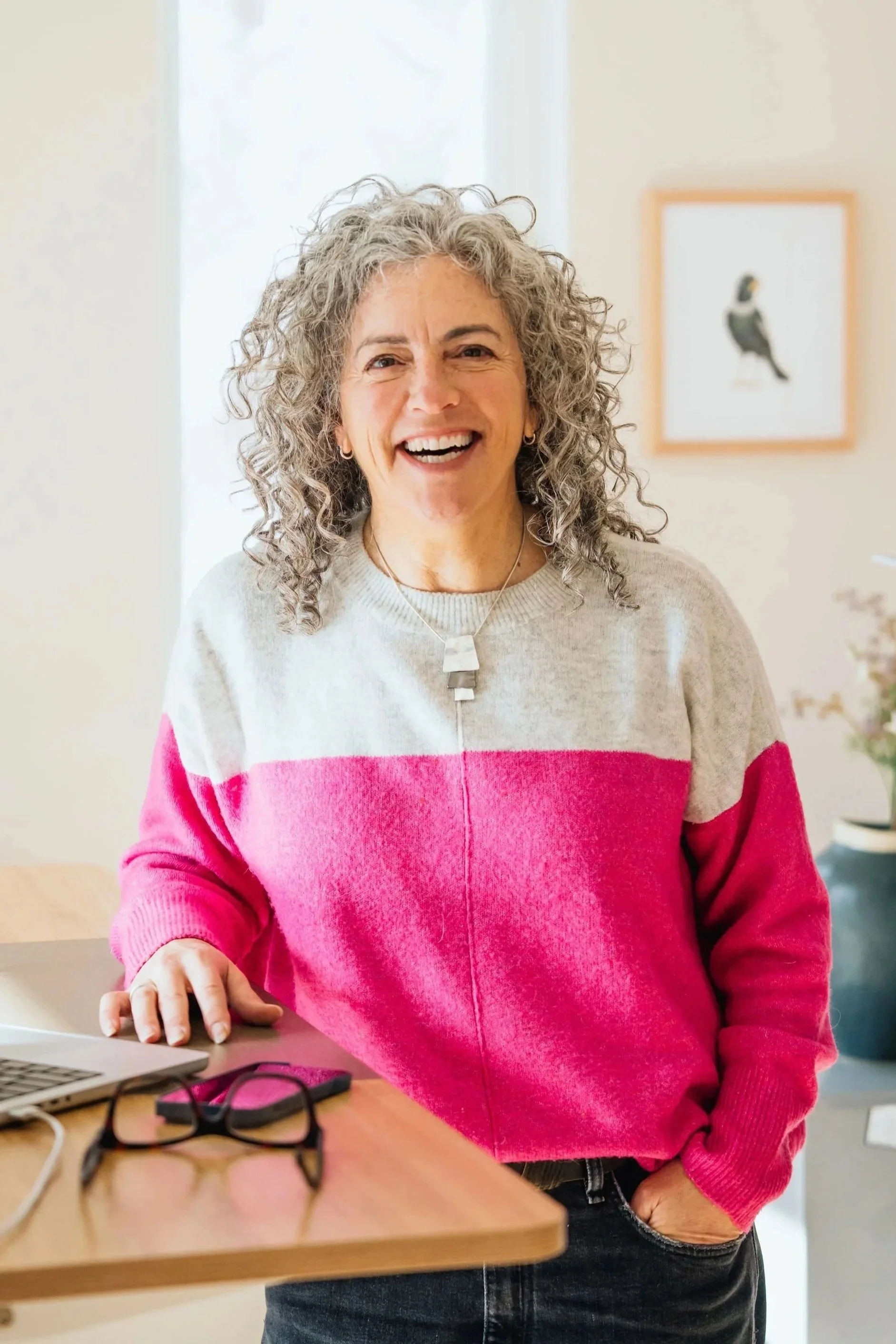 A woman with curly gray hair is smiling and standing in a room with a framed picture of a bird in the background. She is wearing a color-blocked sweater in beige, pink, and red, and has earrings and a pendant necklace. Her right hand is resting on a table with glasses, a laptop, and other items on it.