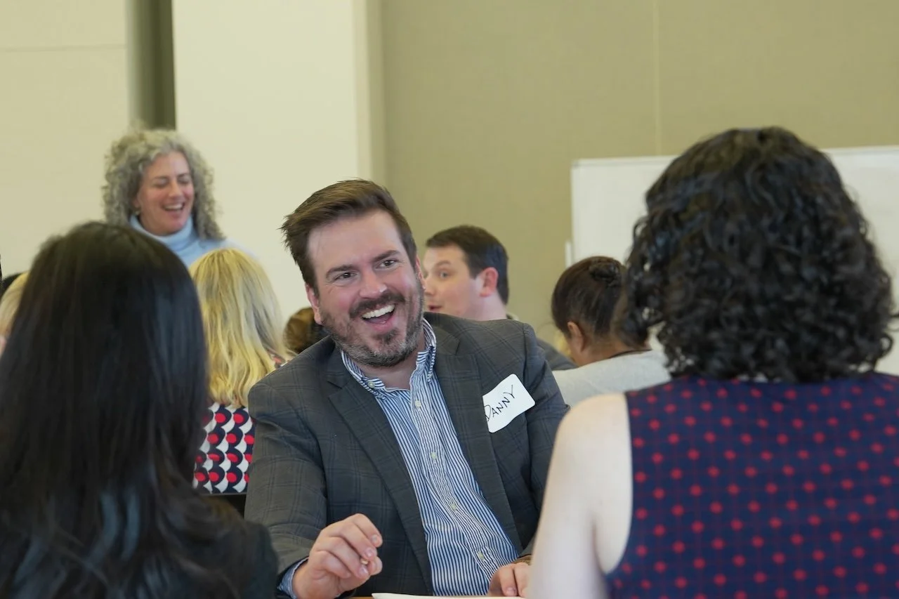 A group of people sitting at tables in a conference or workshop setting, engaging in conversation and smiling.