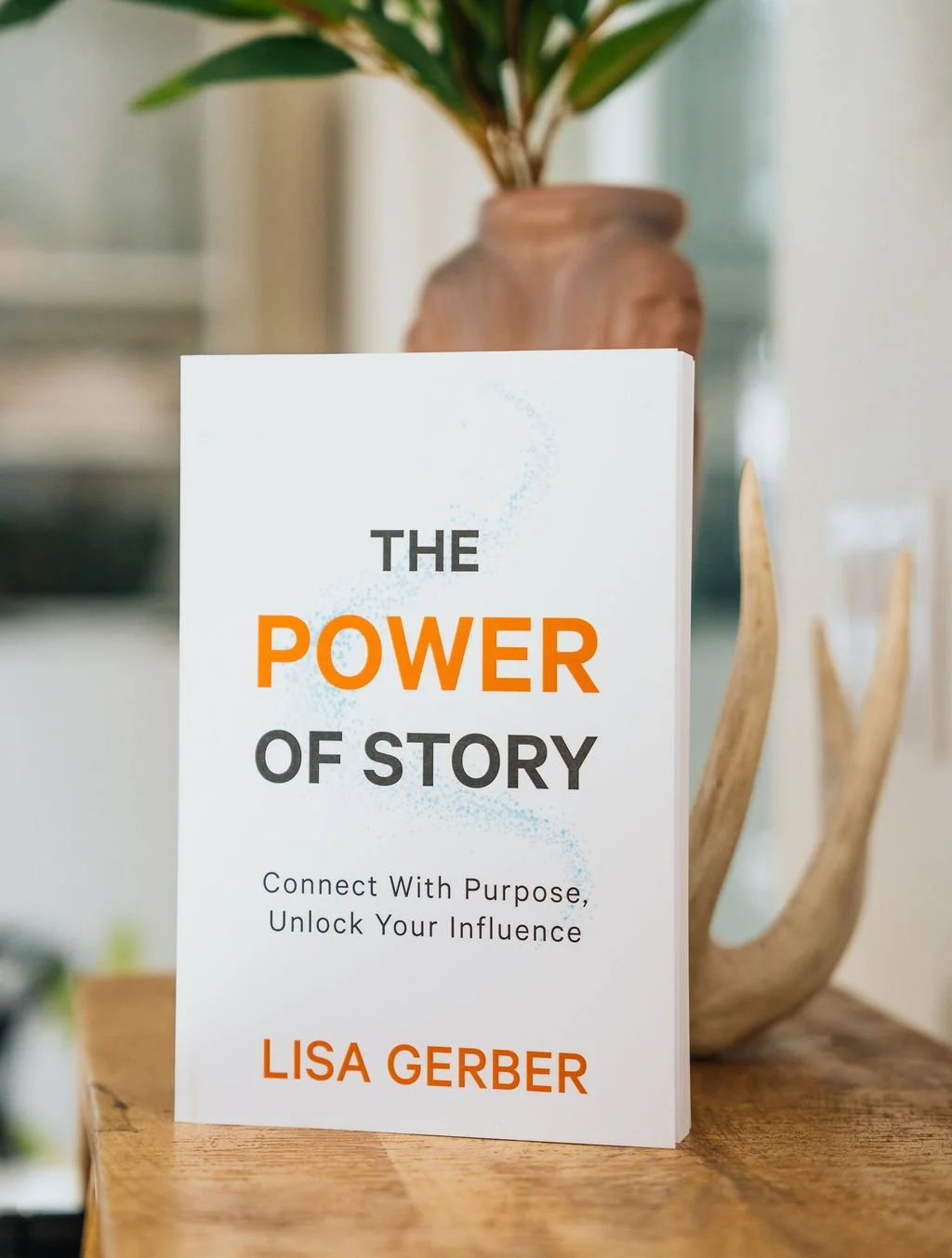Book titled 'The Power of Story' by Lisa Gerber standing on a wooden surface with a ceramic vase containing a leafy plant in the background.