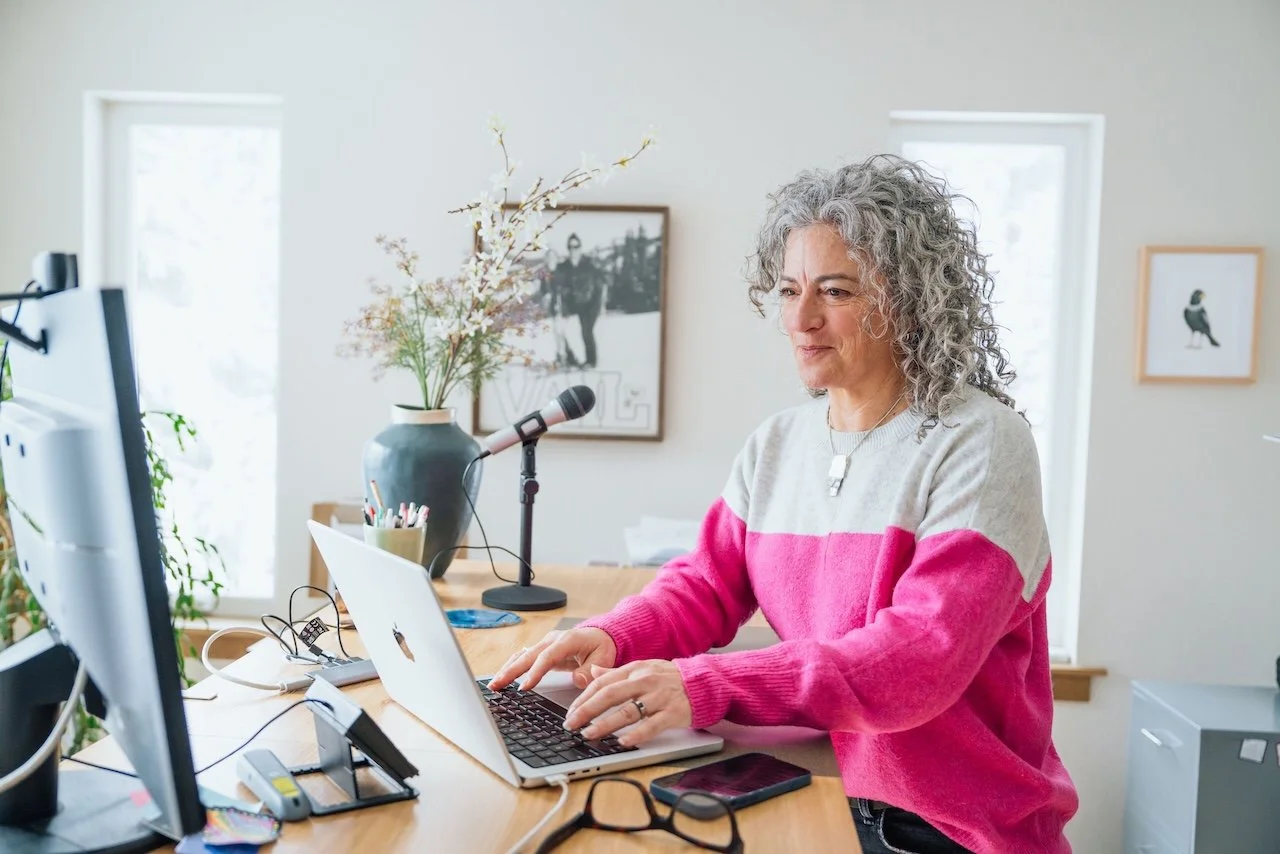 A woman working on a laptop at a wooden desk in a bright room with large windows, a vase with flowers, and framed pictures on the wall.