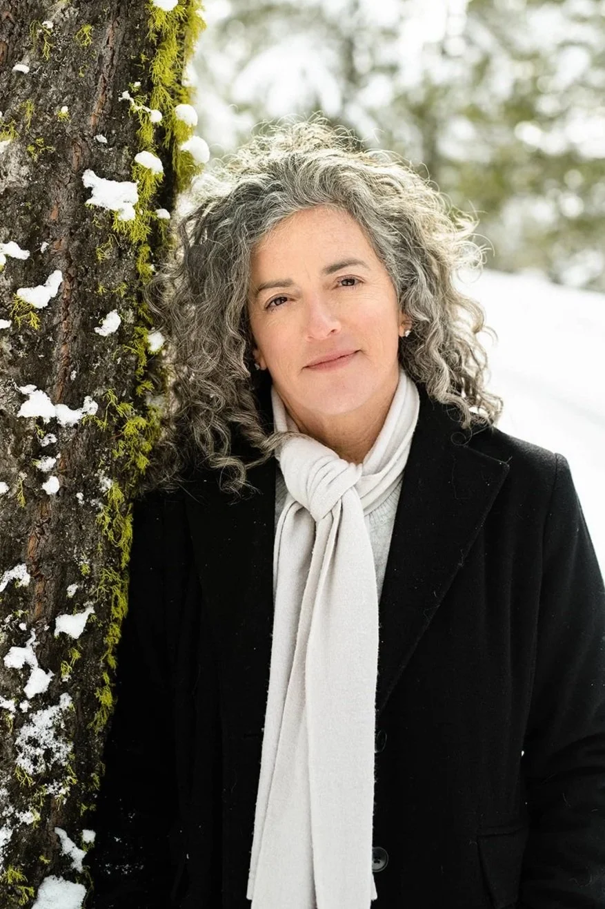 A woman with curly gray hair leaning next to a snow-covered tree trunk in a winter outdoor setting.