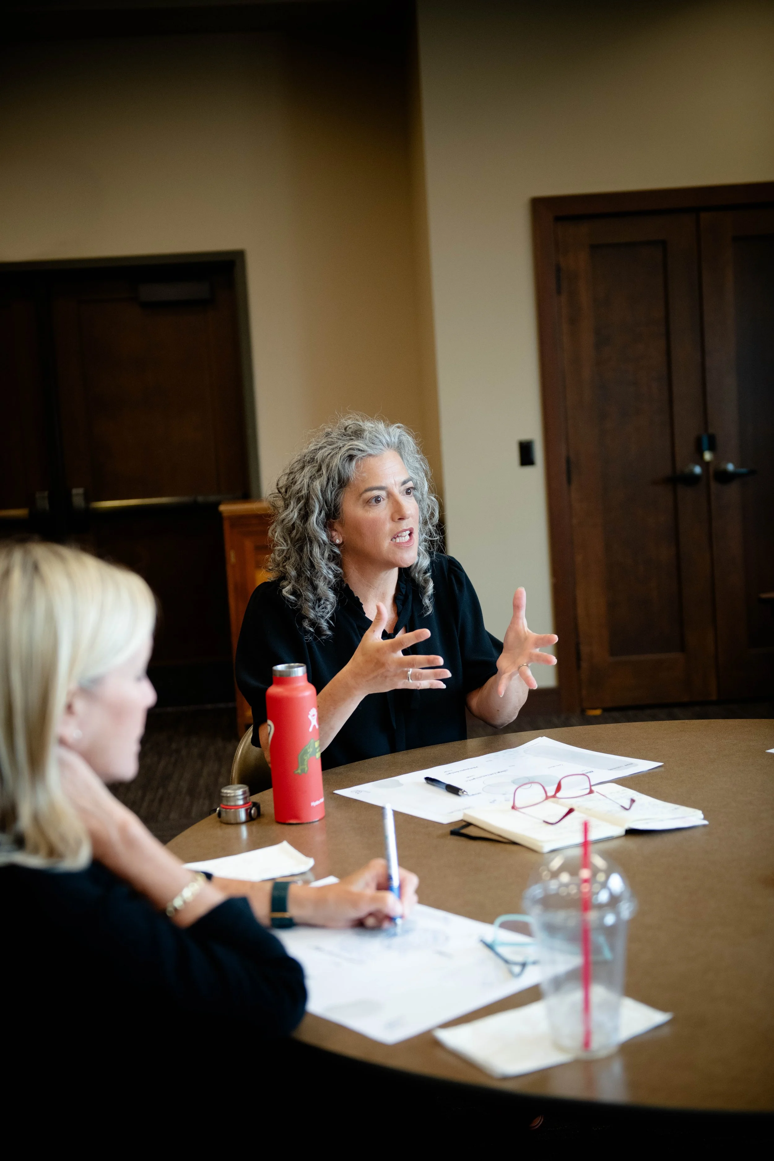 A woman with curly gray hair wearing a black top is speaking and gesturing with her hands during a meeting. There are papers, a red water bottle, and glasses on the table. Other individuals, mostly out of focus, are present. The setting appears to be a conference room with wooden doors and beige walls.