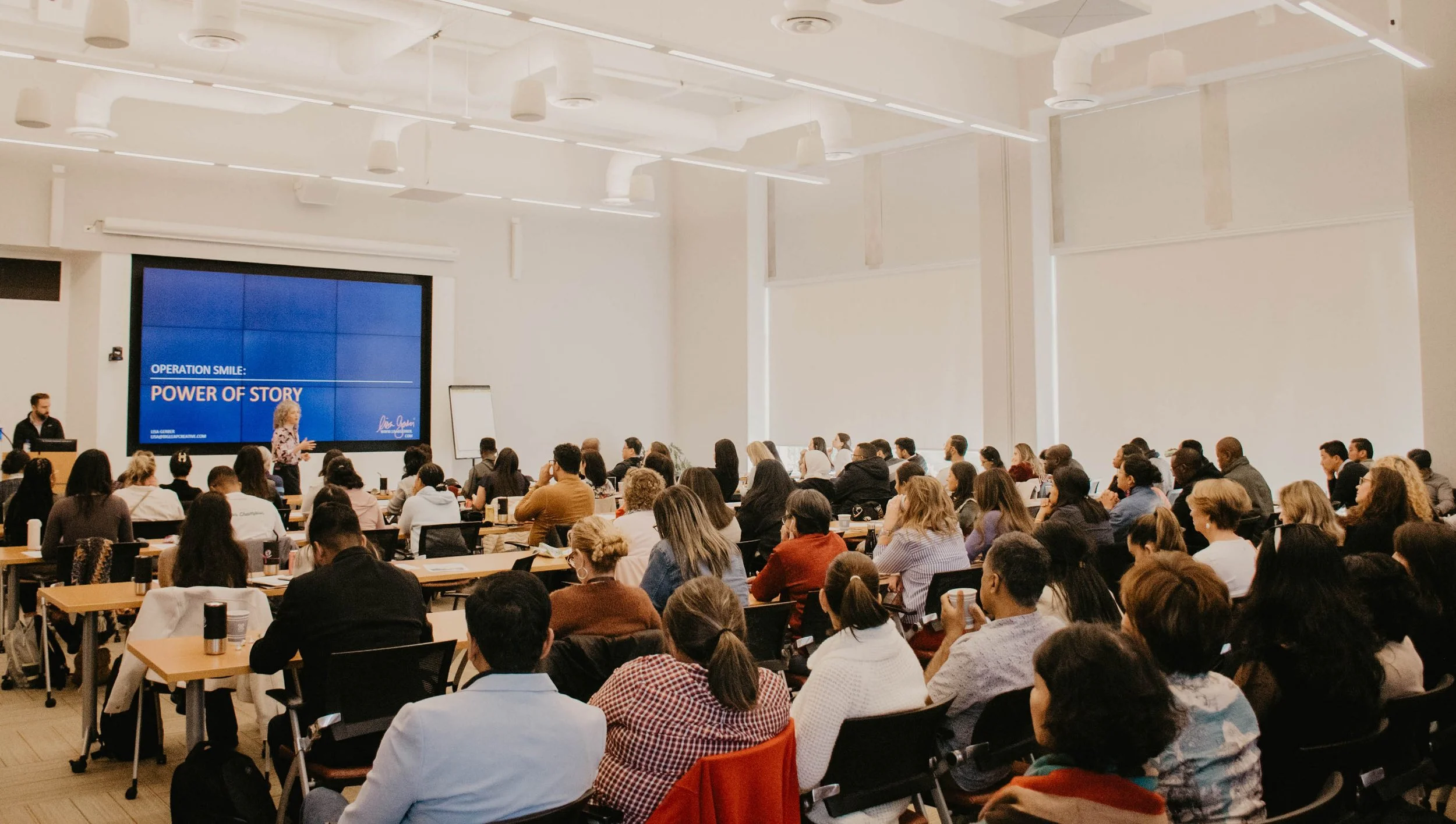 A large conference room filled with many attendees seated at tables, listening to a speaker at the front. A large screen displays 'Operation Smile: Power of Story.' The room has high ceilings with white walls and modern lighting.