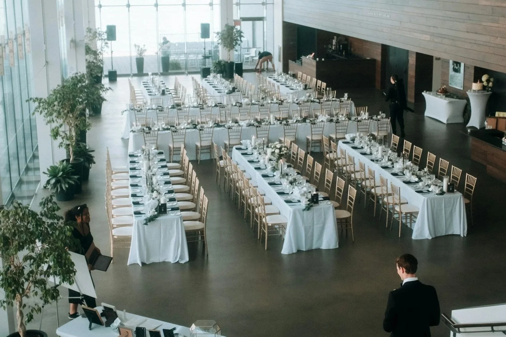 A large banquet hall with long white-draped tables set for an event, decorated with floral centerpieces, surrounded by gold chairs, and staff preparing the space.