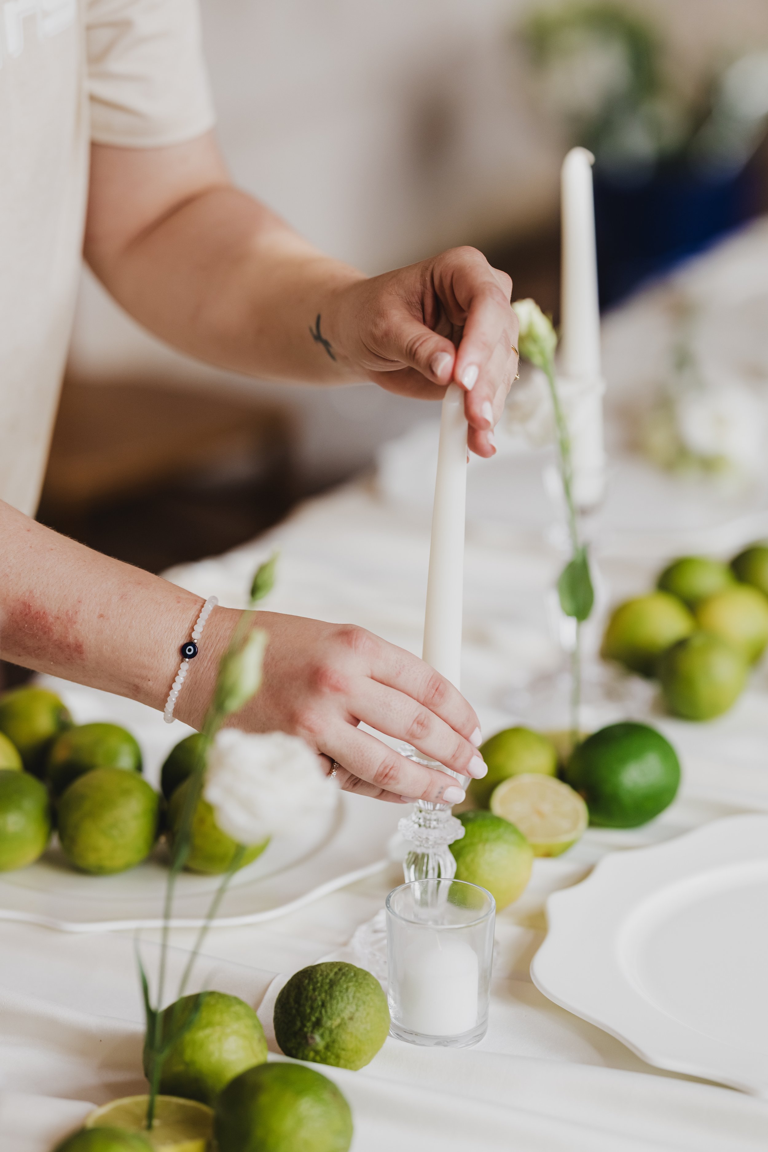 Close-up of a person's hand placing a candle on a table decorated with limes, flowers, and other candles, likely for a celebration or event.