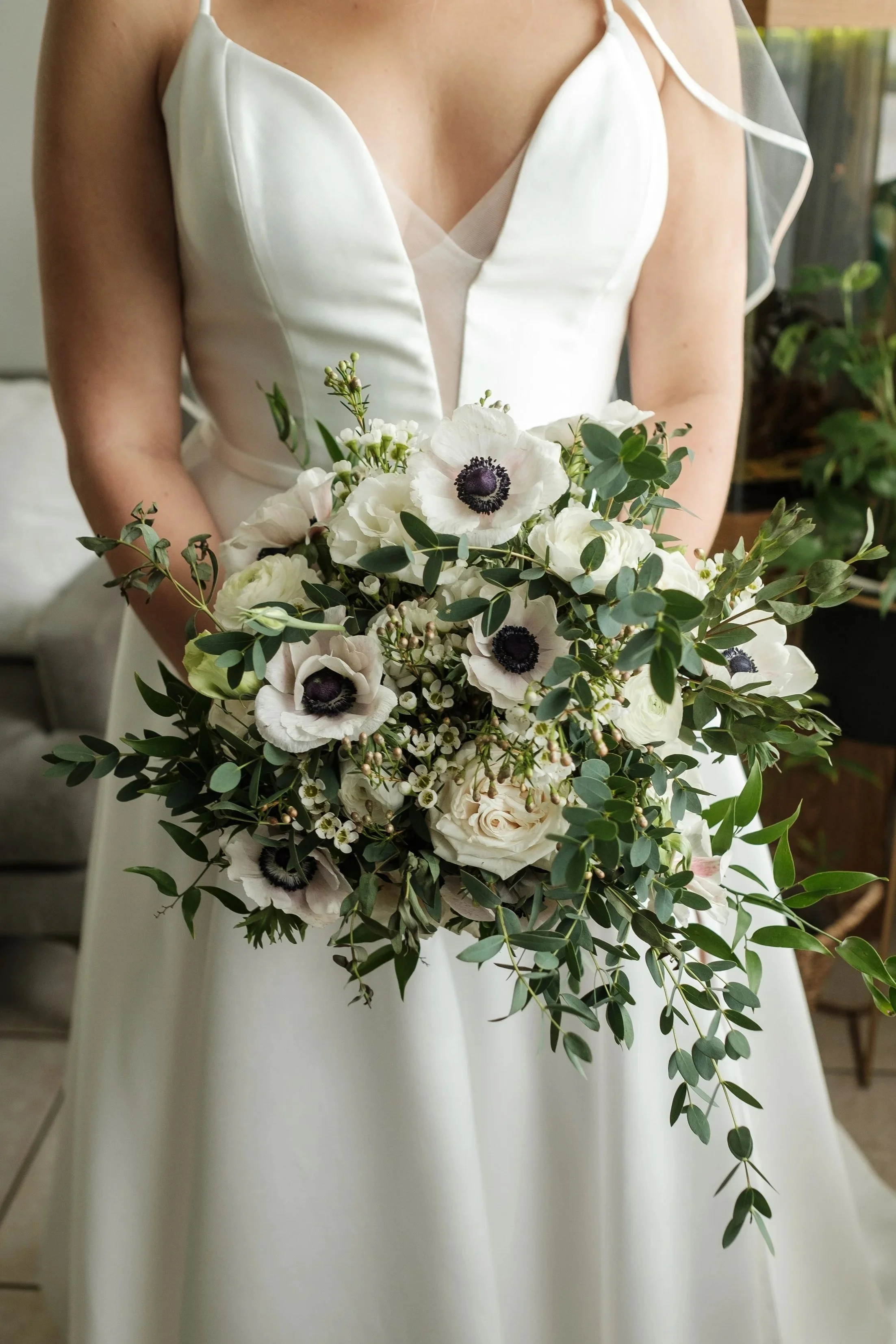 A bride wearing a white satin wedding gown with a V-neckline and sheer overlay, holding a bouquet of white flowers and greenery, standing indoors with plants in the background.