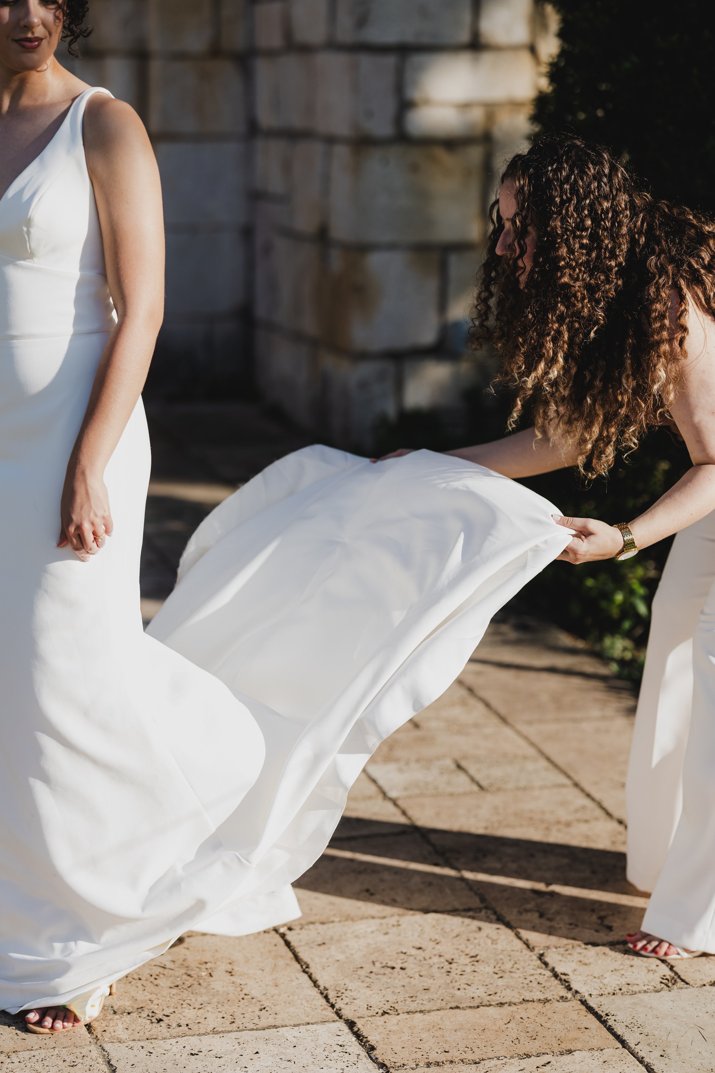 There are three women in white dresses, one of whom is holding the train of another woman's dress, suggesting a wedding or similar formal event. They are standing on a paved area outdoors, with a large stone wall and some greenery in the background.