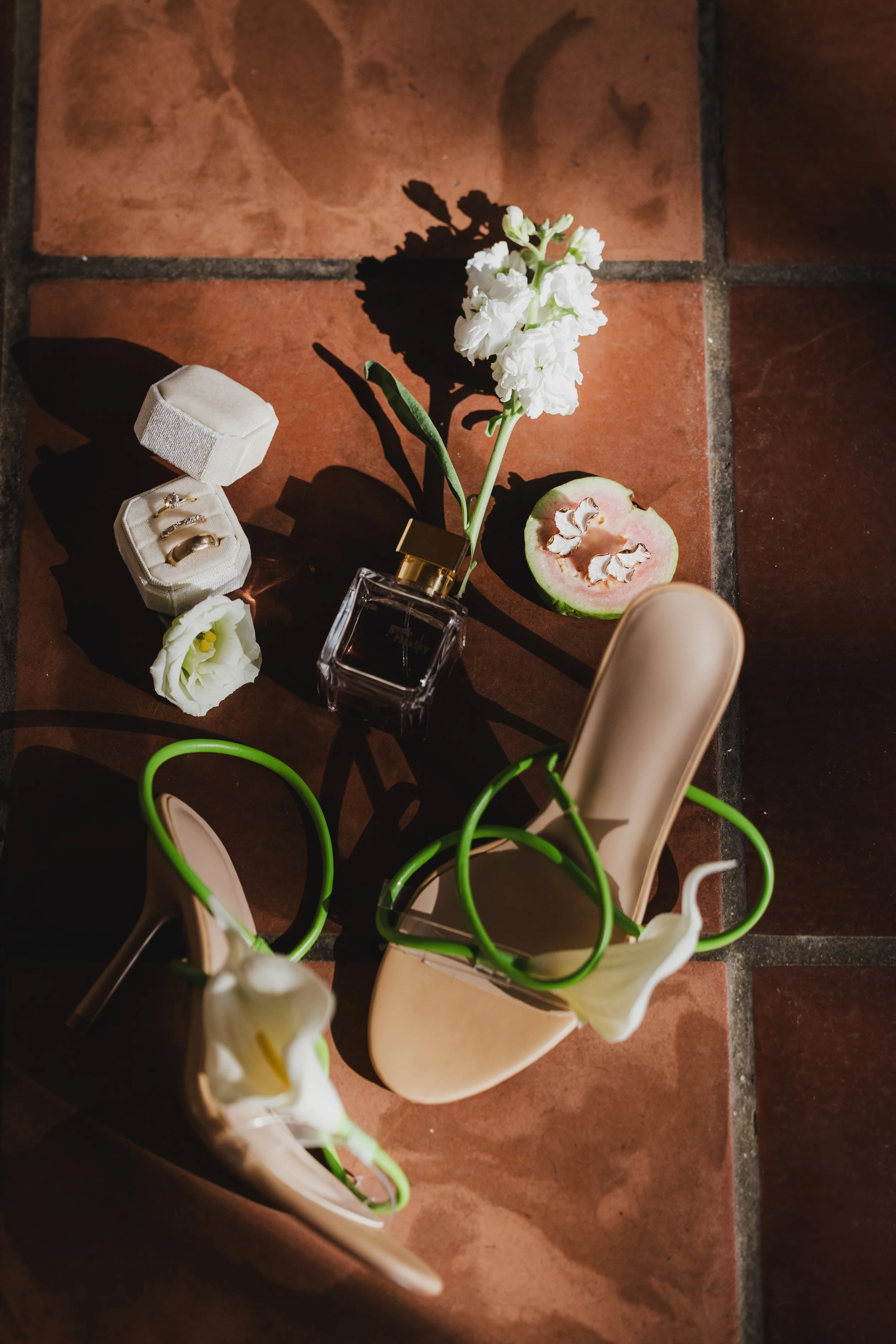 A pair of beige high-heeled shoes with green and white floral accents, a white flower, a small white ring box with three rings inside, a white flower in a small container, a half-cut guava fruit, a black perfume bottle with a gold cap, and a sliced guava with seeds, all arranged on a brown tiled floor.