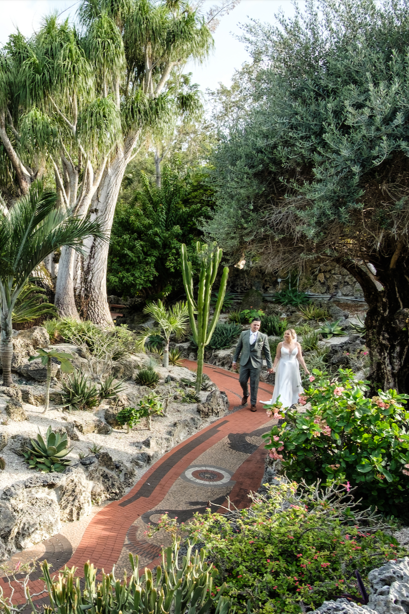 A bride and groom walking hand in hand along a winding garden path surrounded by lush trees and plants.
