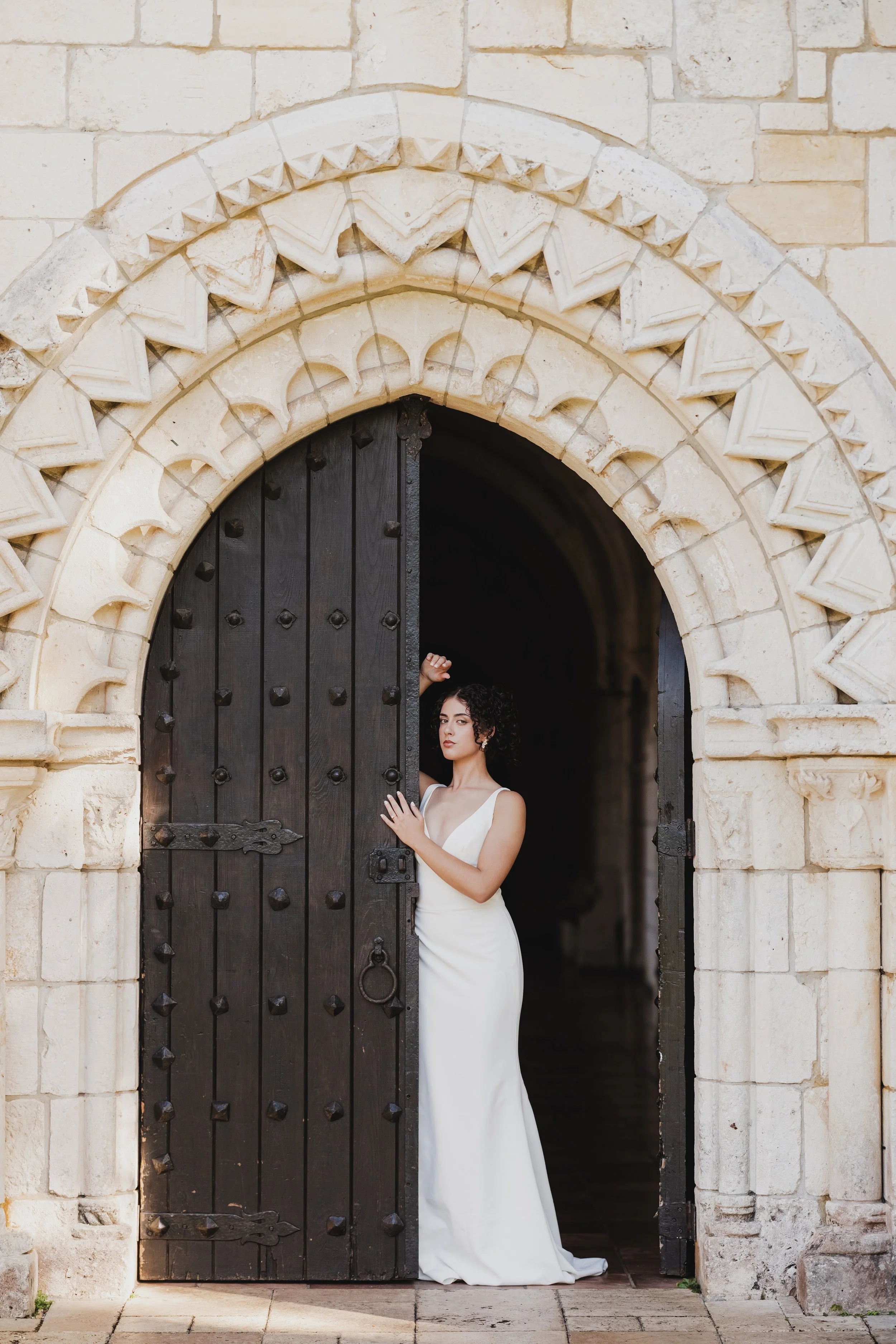 A woman in a white wedding dress stands in the doorway of an old stone building, partially obscured by a large, dark wooden door.
