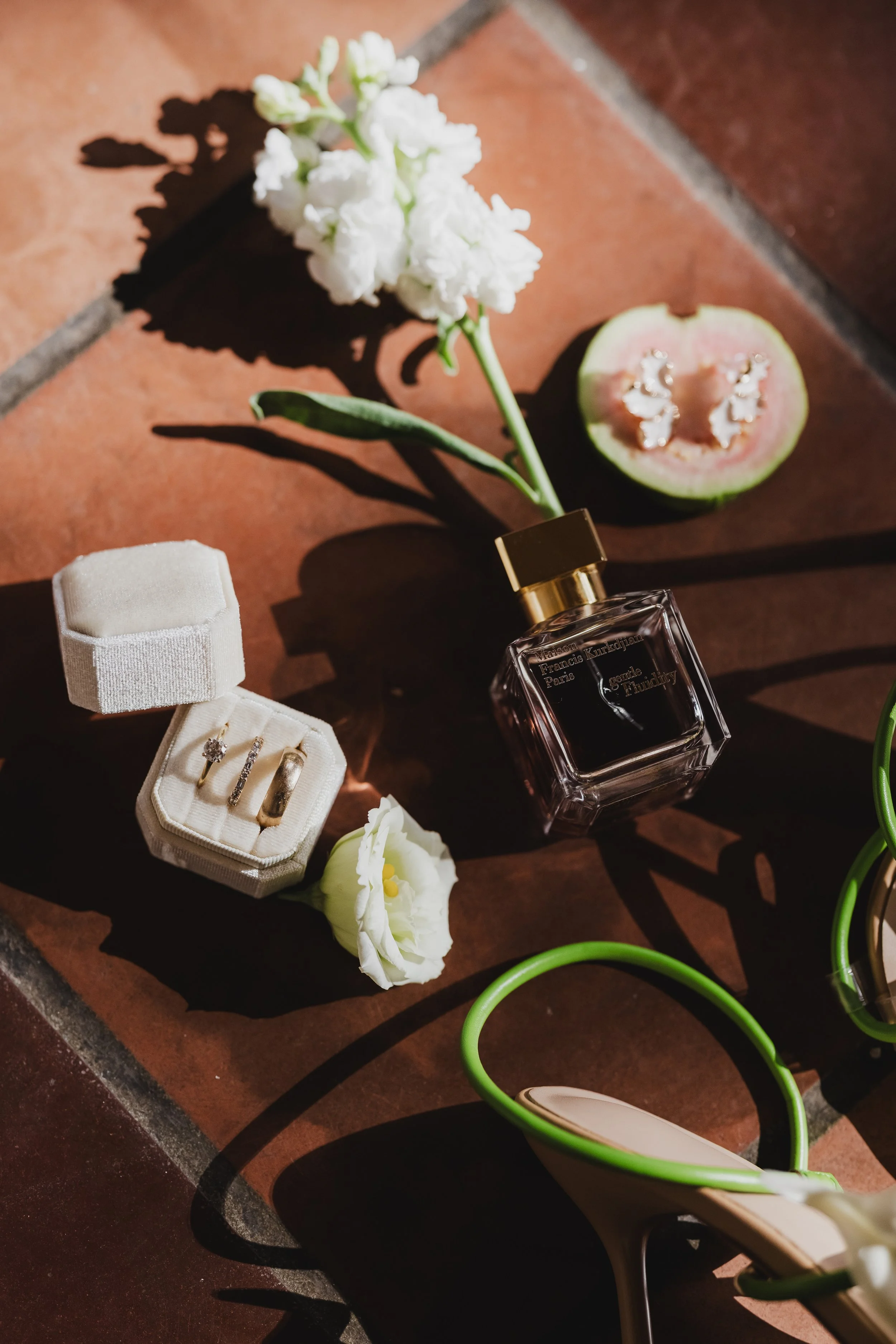 A white flower, a half-cut guava fruit, a jewelry box with three rings, a white and green object, and a small white flower, all arranged on a terracotta tiled surface.
