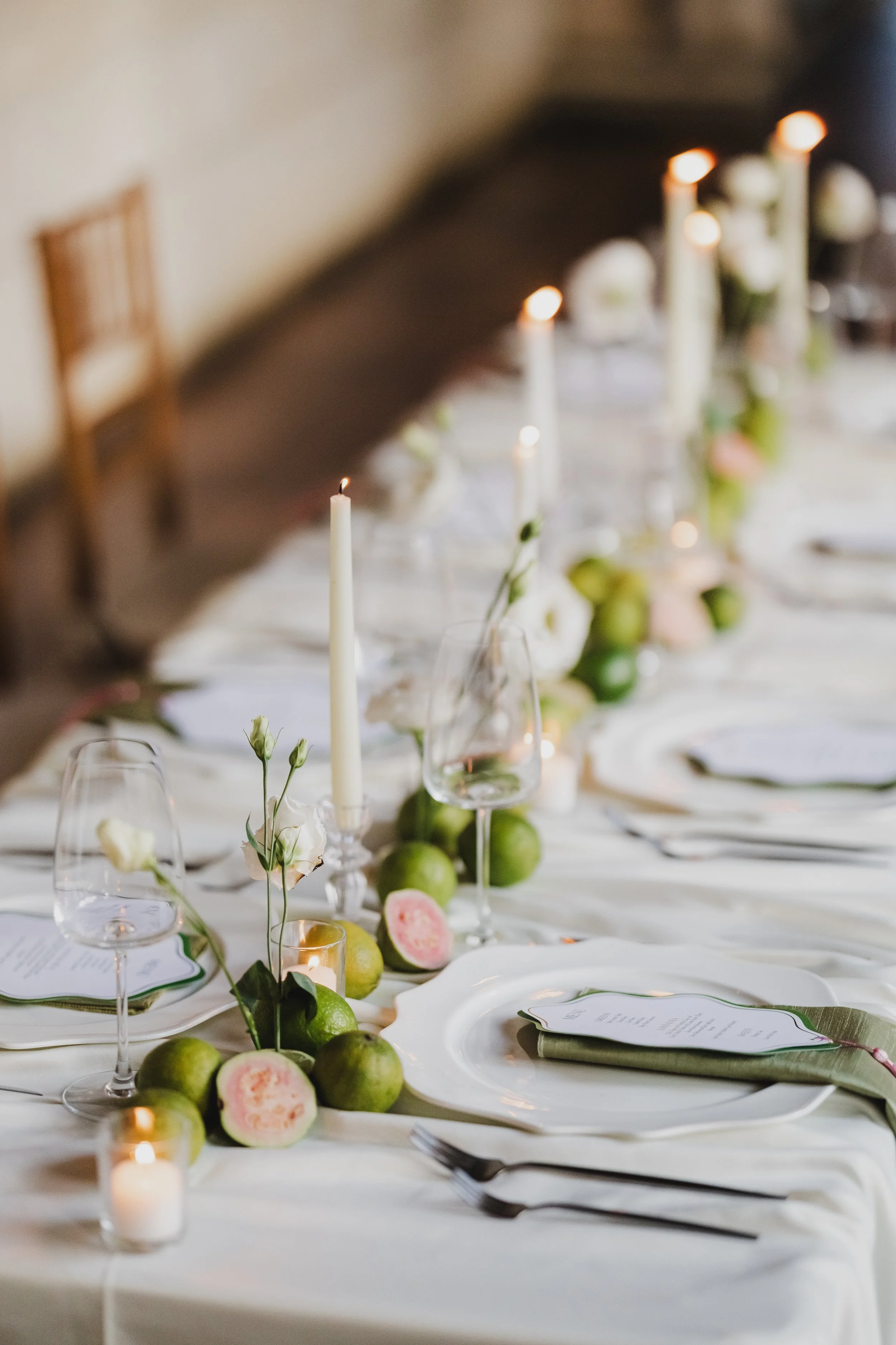 A beautifully set banquet table with white tablecloth, elegant white and pink flowers, green apples, lit candles, wine glasses, and plates with menus.